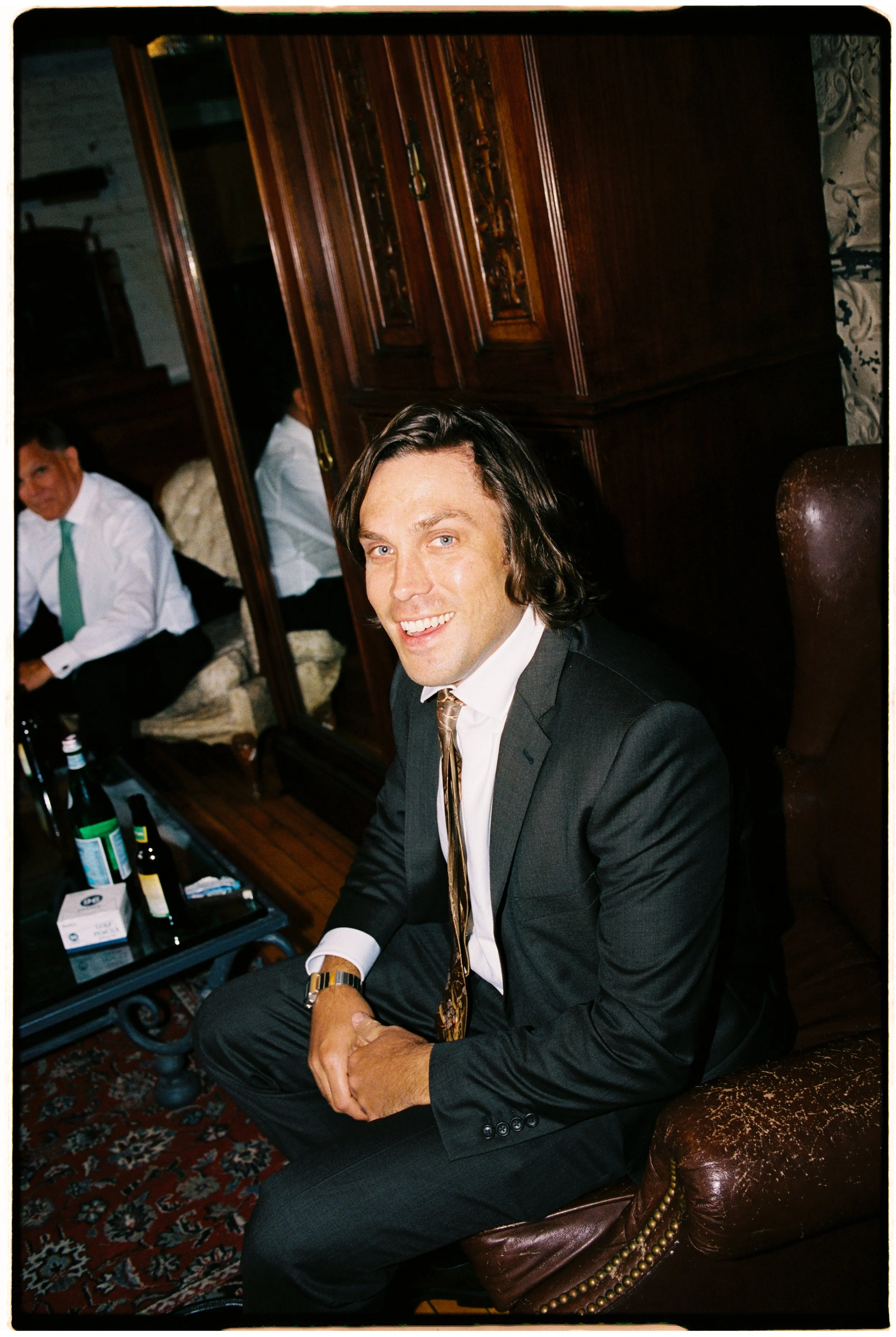 A young man with long dark hair and blue eyes dressed in a black suit, white shirt, and tie, sitting on a leather chair in a room with wooden furniture, a patterned rug, and a mirror, smiling at the camera.