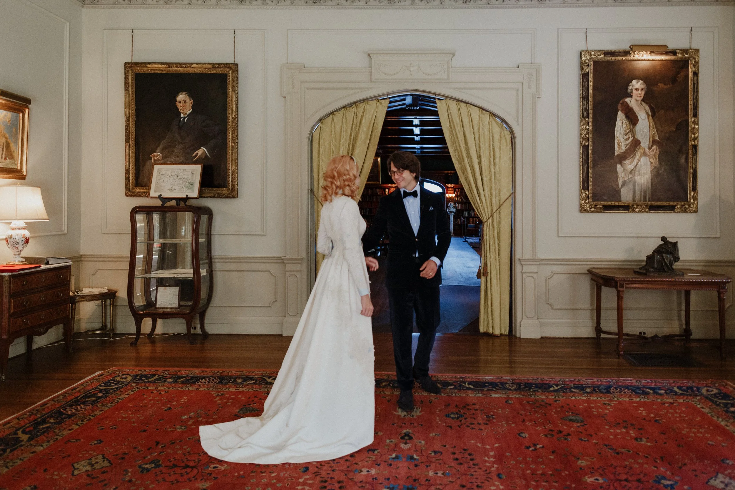 A bride and groom hold hands and smile at each other in a formal room with portraits on the walls and a patterned red carpet.