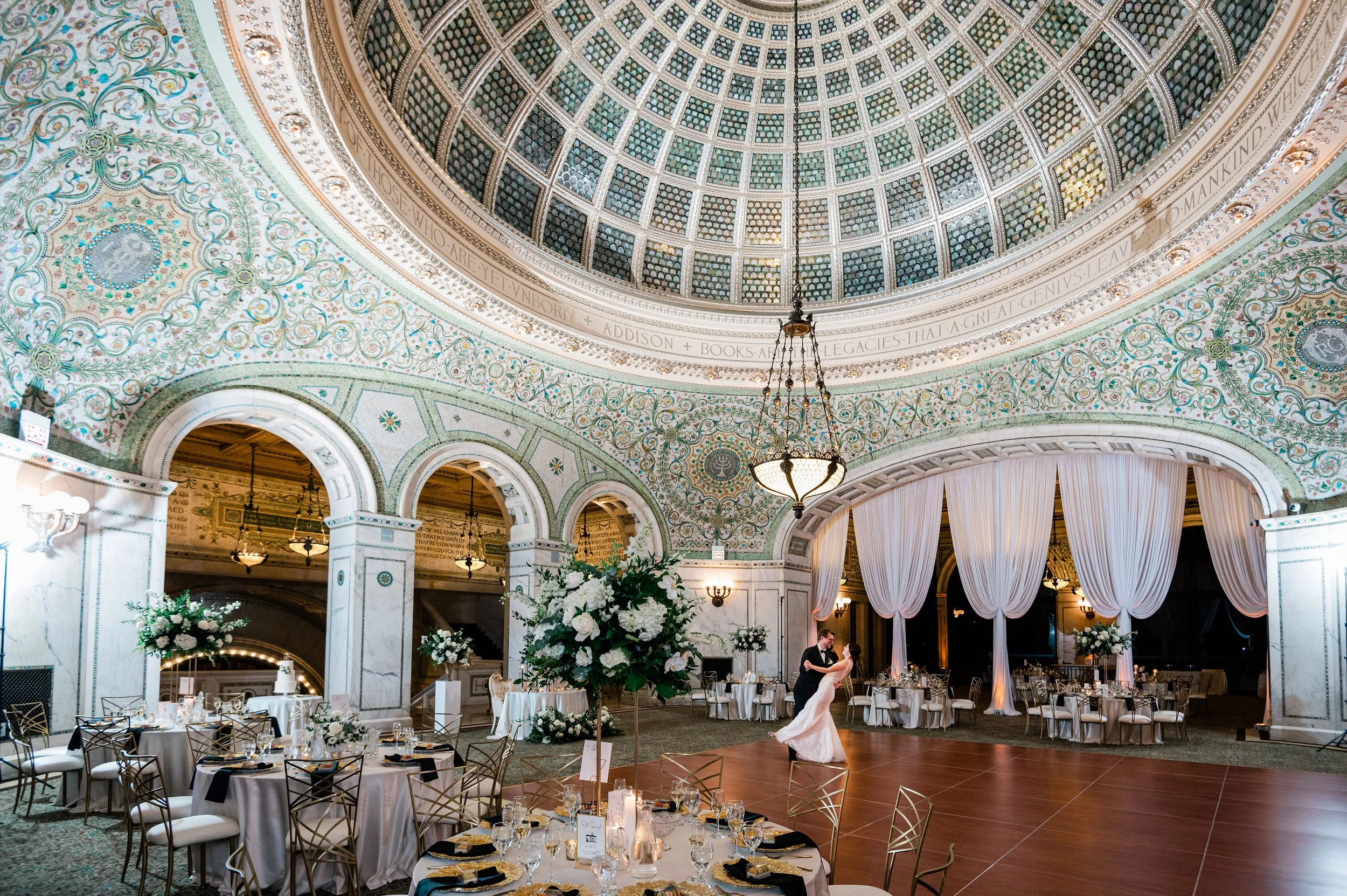 Elegant wedding reception hall with ornate domed ceiling, floral arrangements, round tables set for dining, and a couple dancing.
