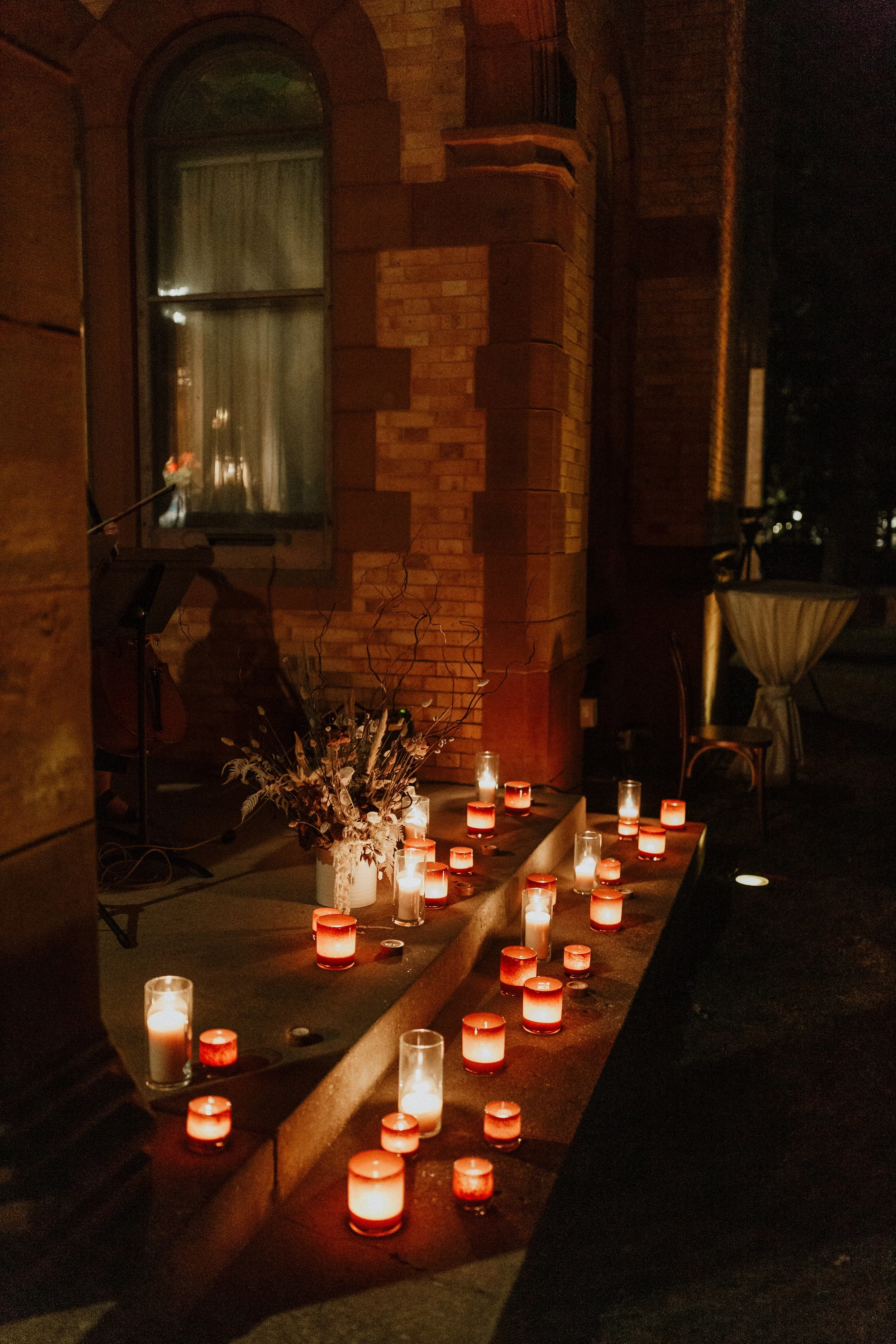 Nighttime scene of a windowsill decorated with numerous candles in glass holders, with a potted plant and a flower arrangement. The scene includes a brick building, a window with curtains, and some chairs and tables, creating a cozy, warm atmosphere.