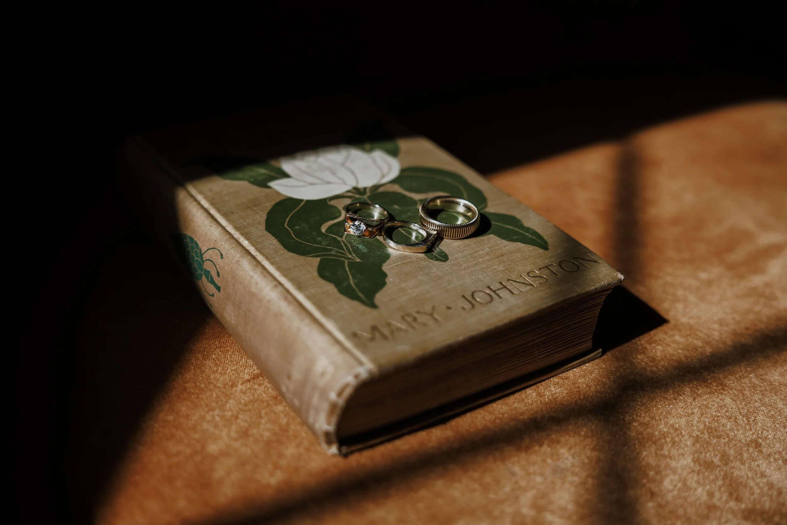 A hardcover book with the title partially visible, placed on a textured surface. Three rings rest on top of the book, illuminated by a focused light in a dark setting.