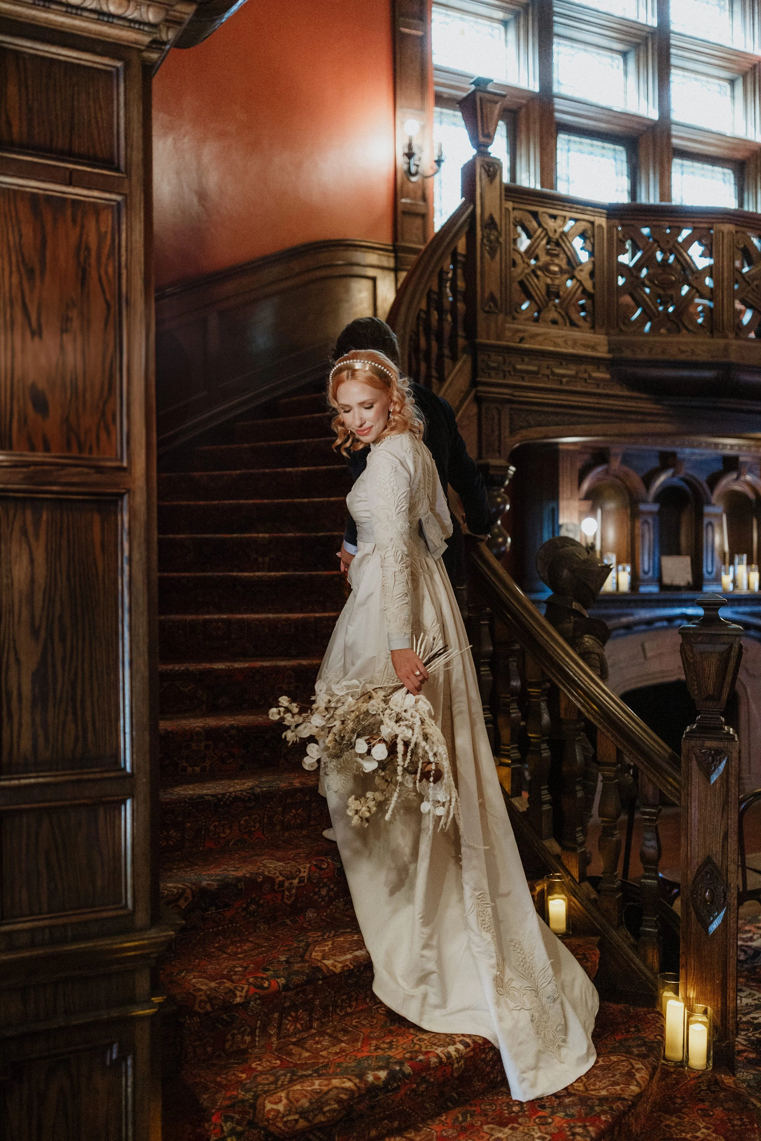 A bride in a white wedding gown holding a bouquet stands on a staircase inside a historic building with wood paneling and stained glass windows.