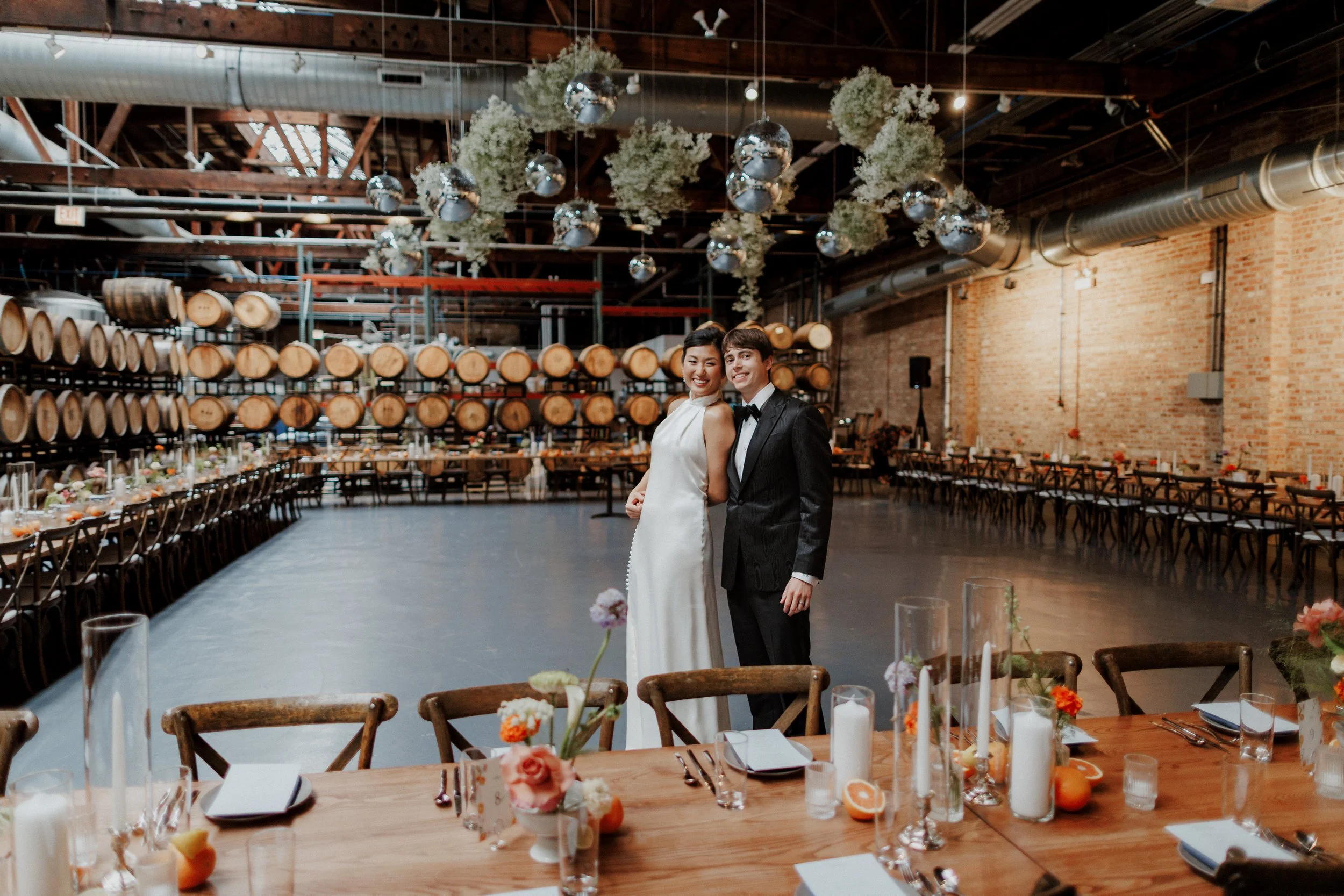 A bride and groom standing together at a wedding reception in an industrial-style venue with exposed brick walls, wooden barrels, and hanging greenery and mirrors.