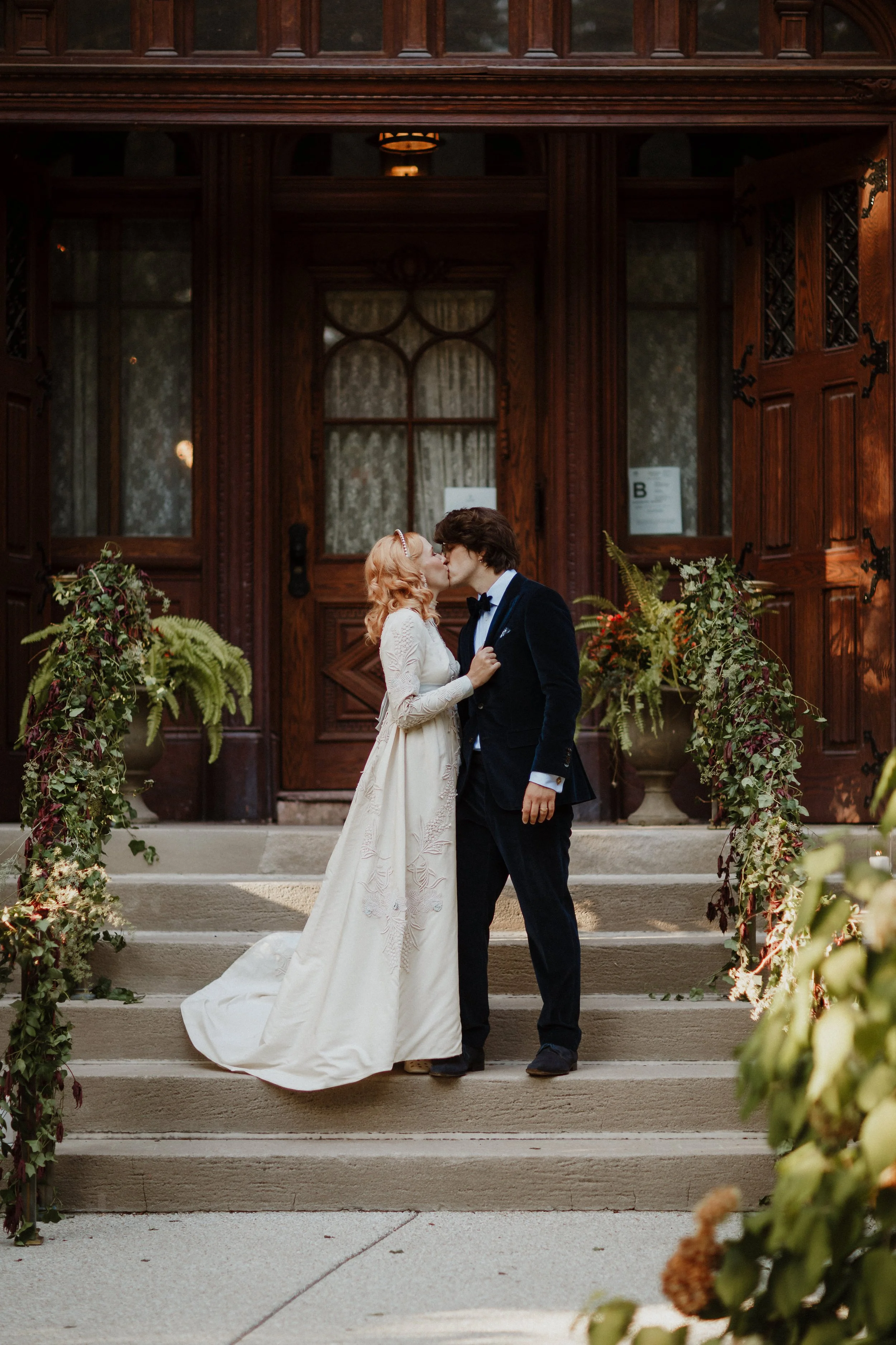 A couple sharing a kiss on wedding day steps, surrounded by greenery and flower arrangements, with a dark wood door in the background.