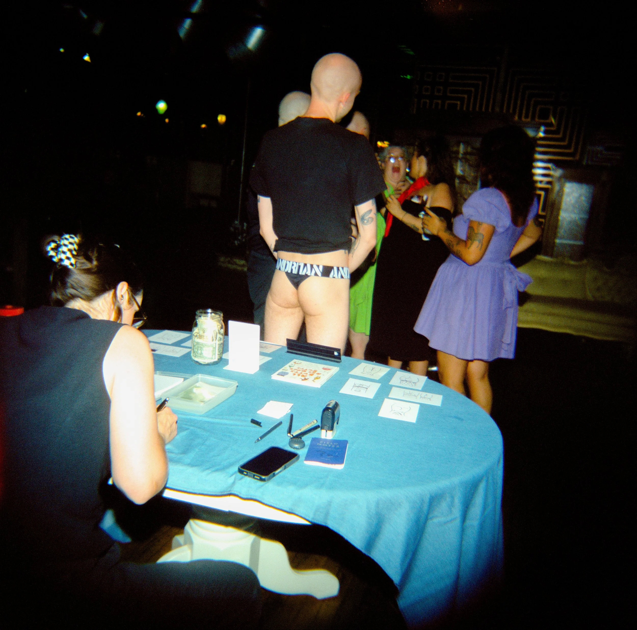 People gathered around a table displaying artwork and photographs at an event, with some engaged in conversation.