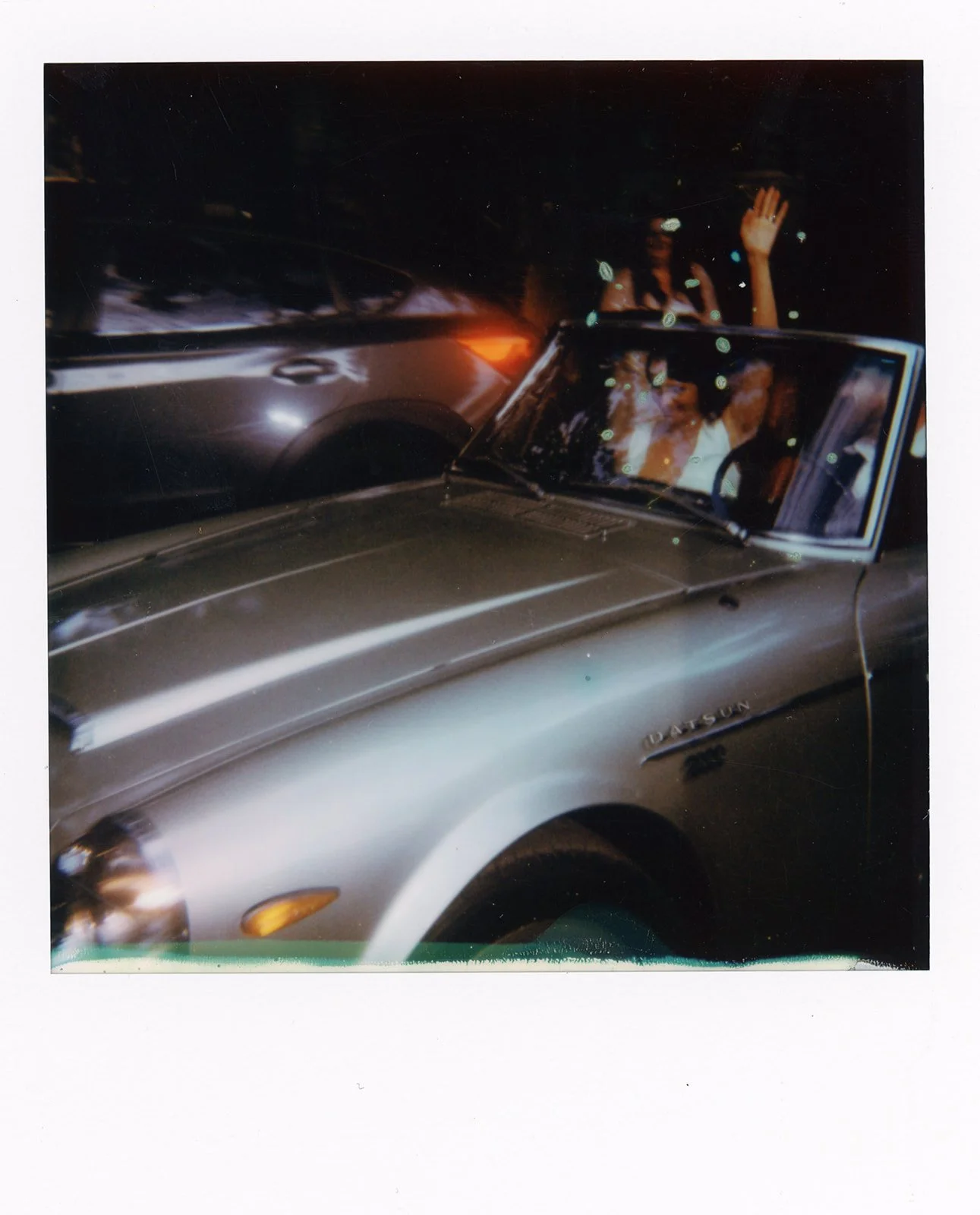 Nighttime scene of a silver vintage Oldsmobile with people inside, one woman waving, windshield reflections, and nearby parked cars.