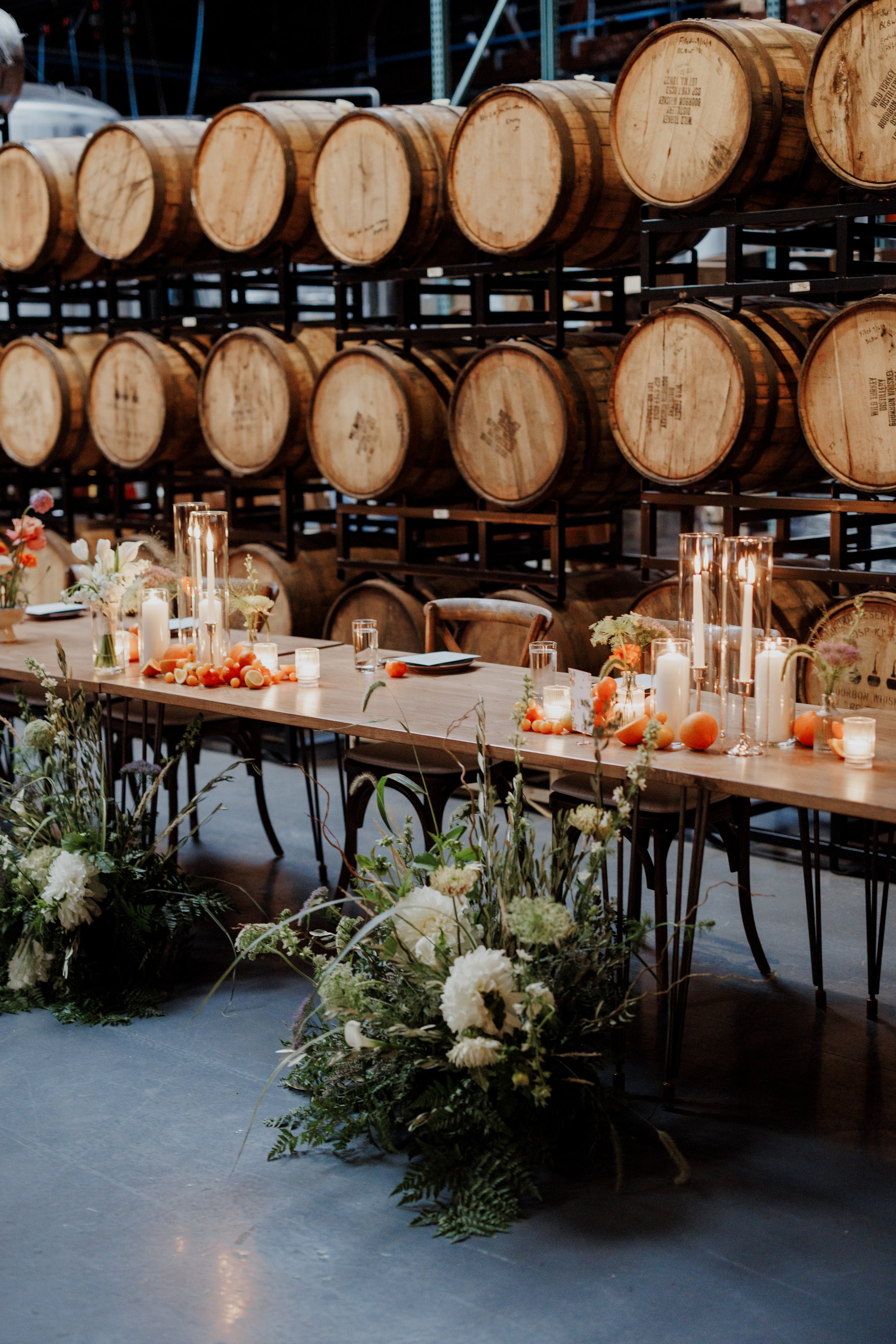 A table decorated with candles, flowers, and oranges, with a backdrop of stacked wooden barrels.