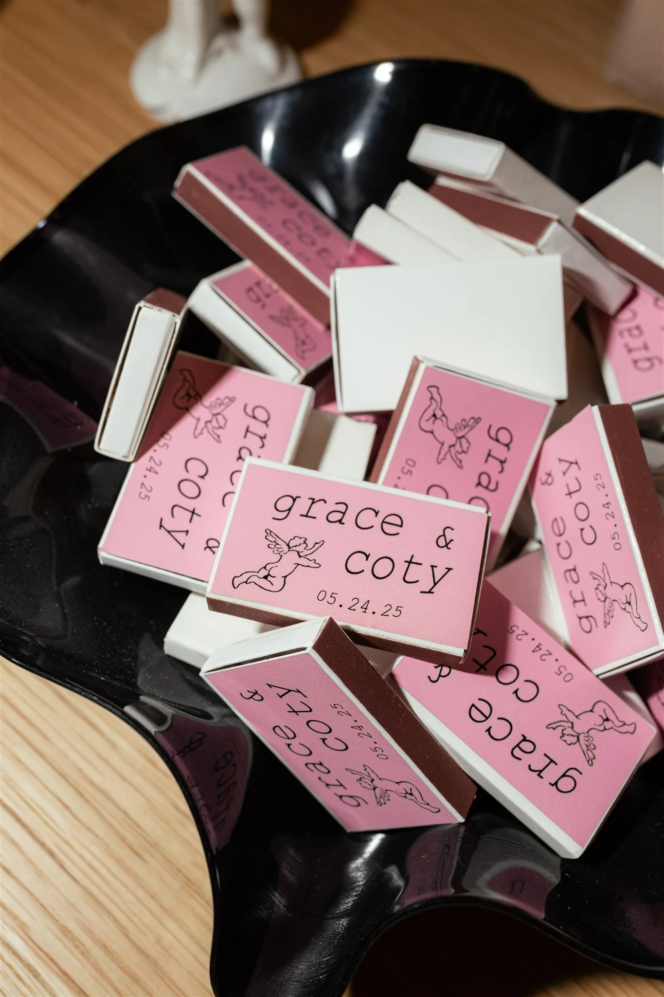 Pink and white matchboxes in a black bowl with a unicorn illustration and date 05.24.25 printed on the boxes, on a wooden surface.