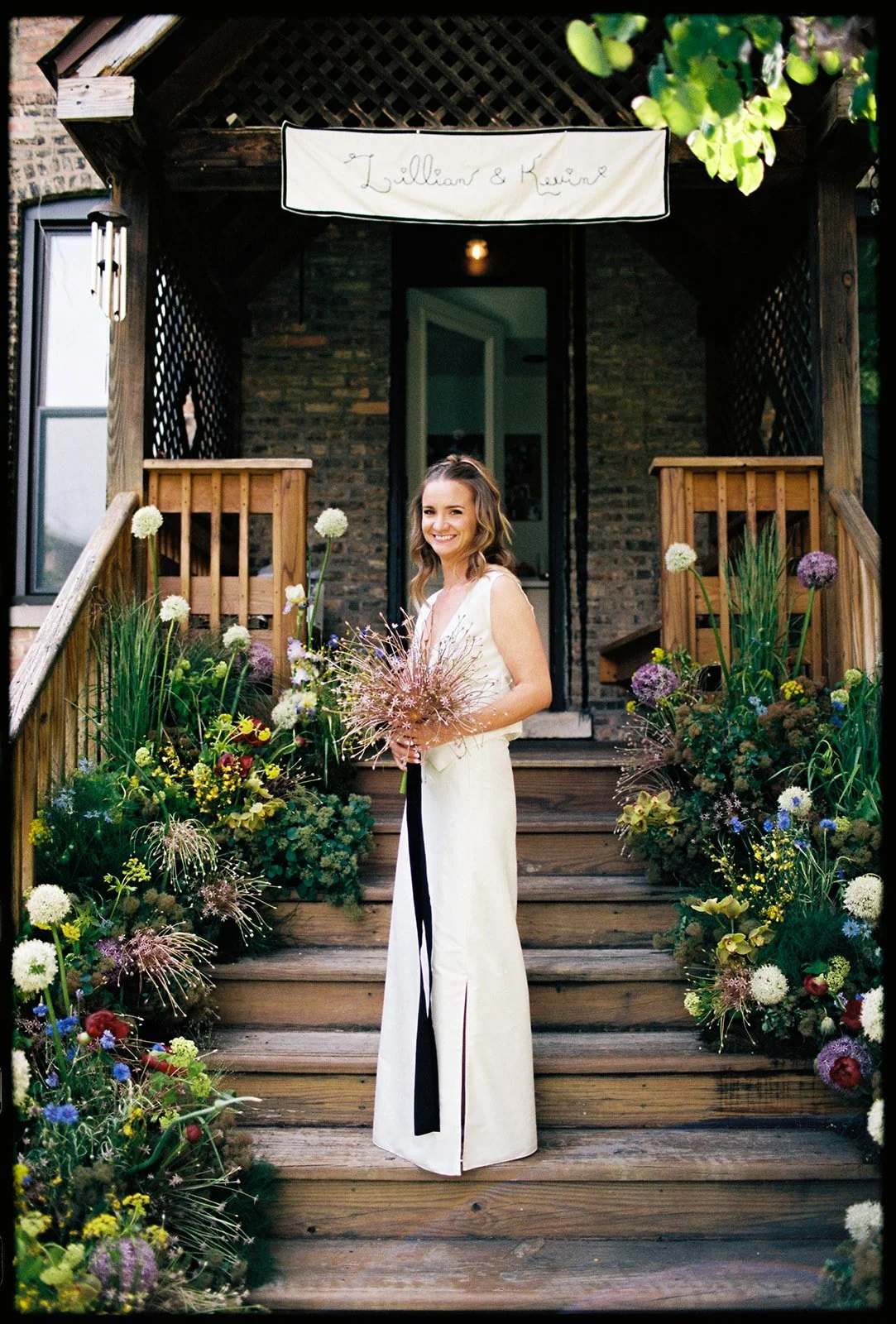 A woman in a white dress standing on wooden stairs decorated with colorful flowers, holding a bouquet, smiling in front of a brick house entrance with a sign that reads 'Lillian & Kevin'.
