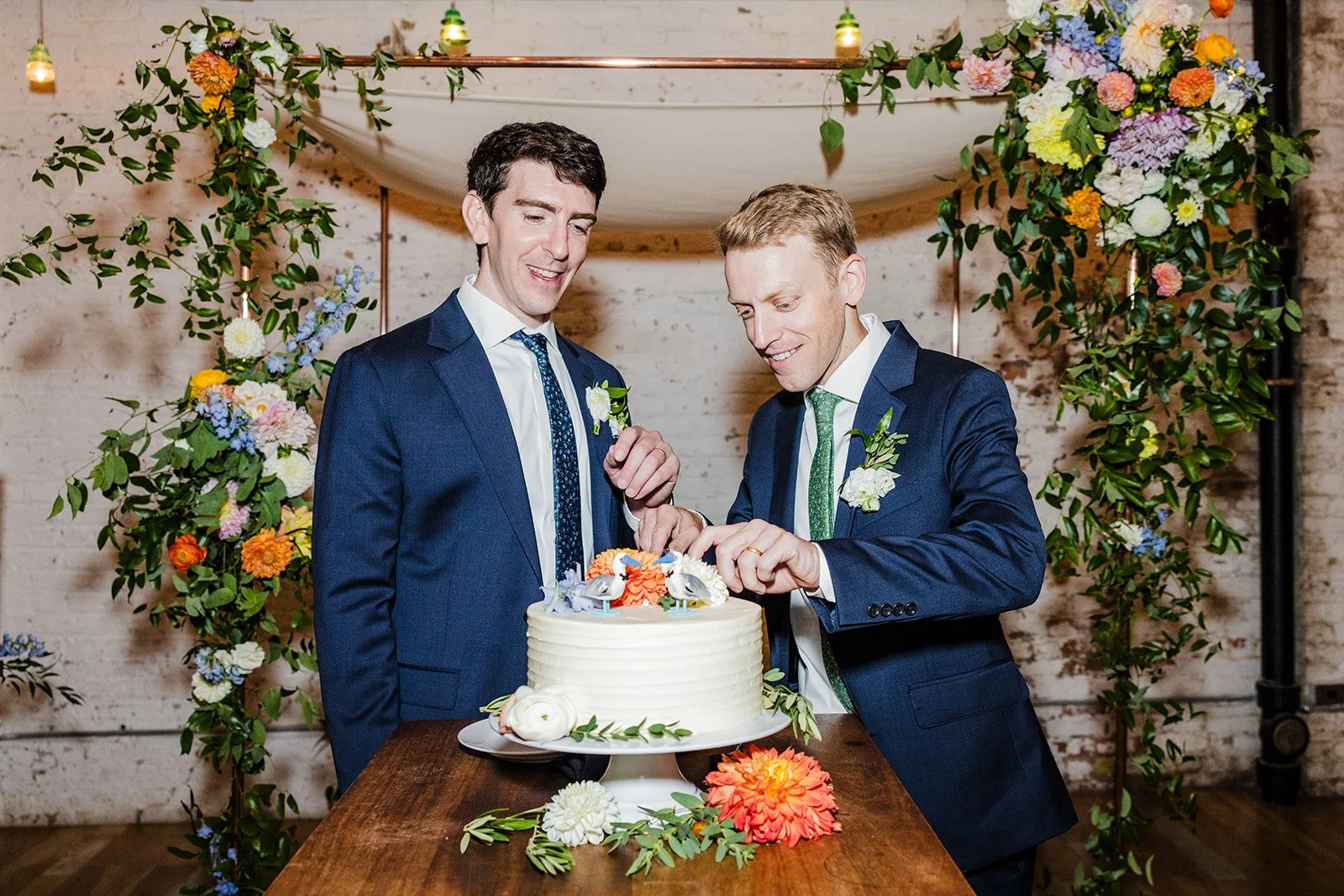 Two men in suits cutting a wedding cake decorated with flowers during a celebration.