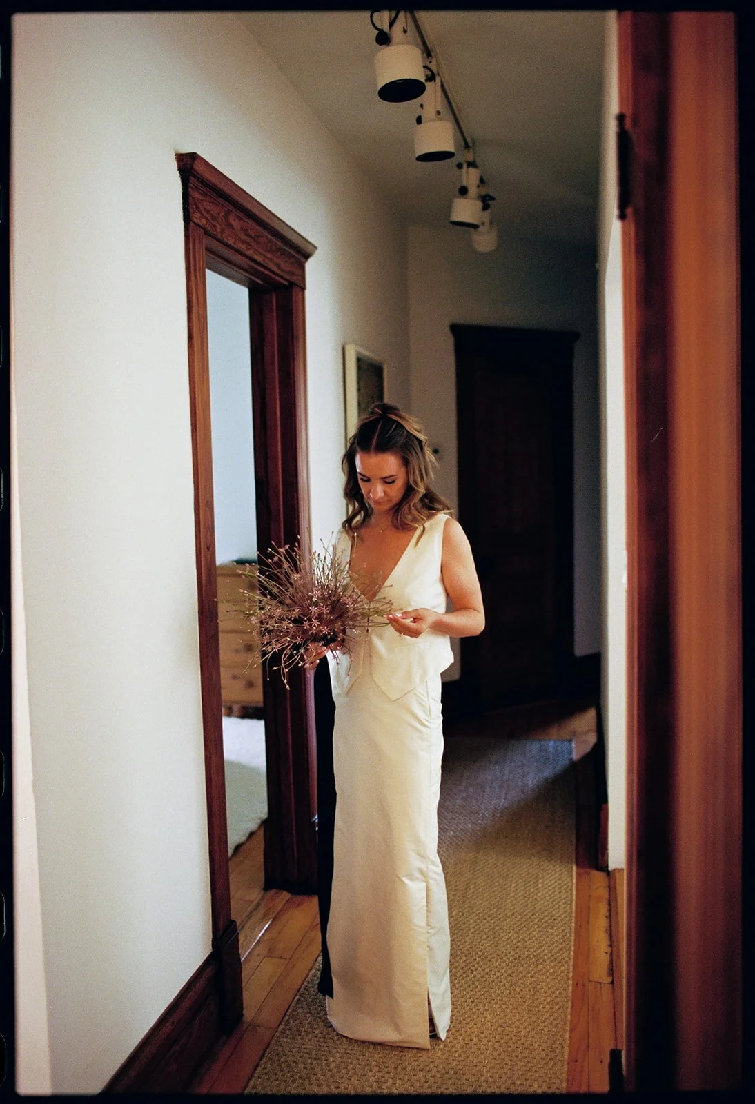 A woman in a white dress holding a bouquet of pink and purple flowers standing in a hallway with wood trim and a mirror.