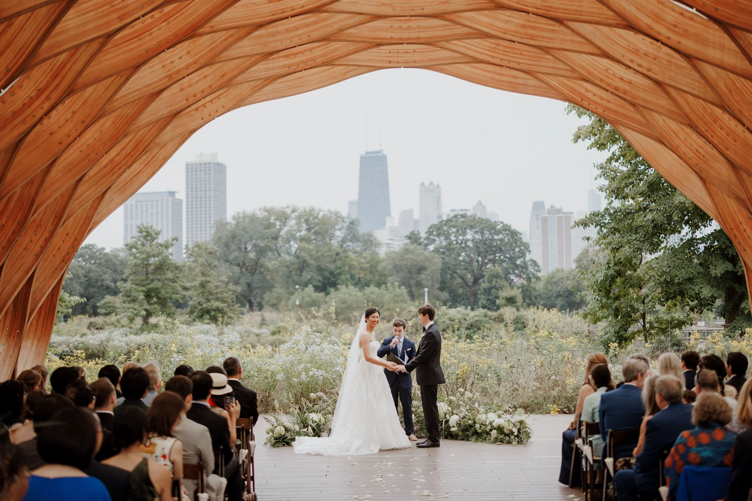 A wedding ceremony taking place outdoors under a wooden arch with the city skyline in the background. The bride and groom are exchanging vows while officiants and guests are present.