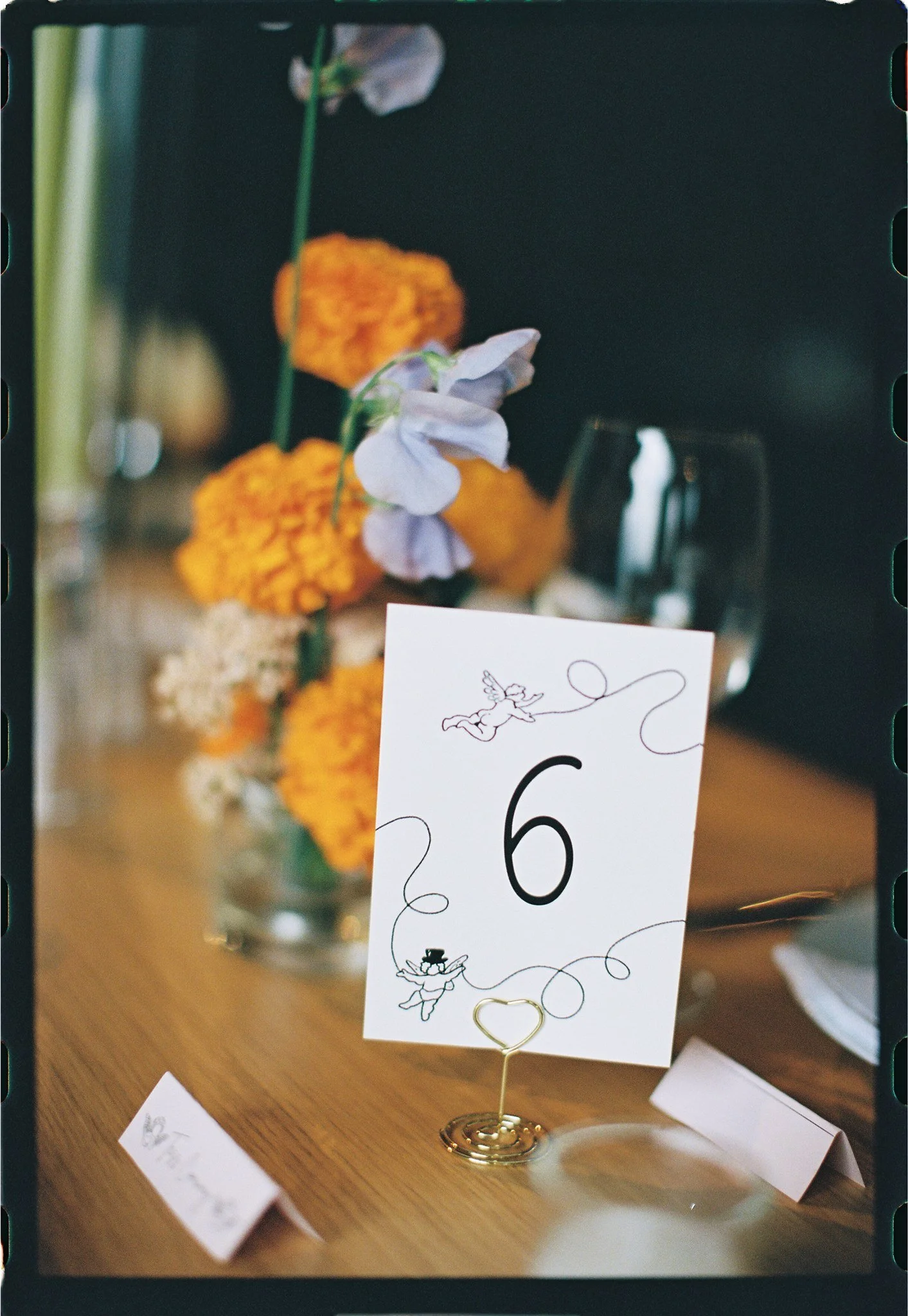 A table displaying a wedding or event table marker with the number 6, decorated with a whimsical illustration of a dancing couple and a unicorn. The table marker is in a gold spiral holder. In the background, there are blurred flowers including white
