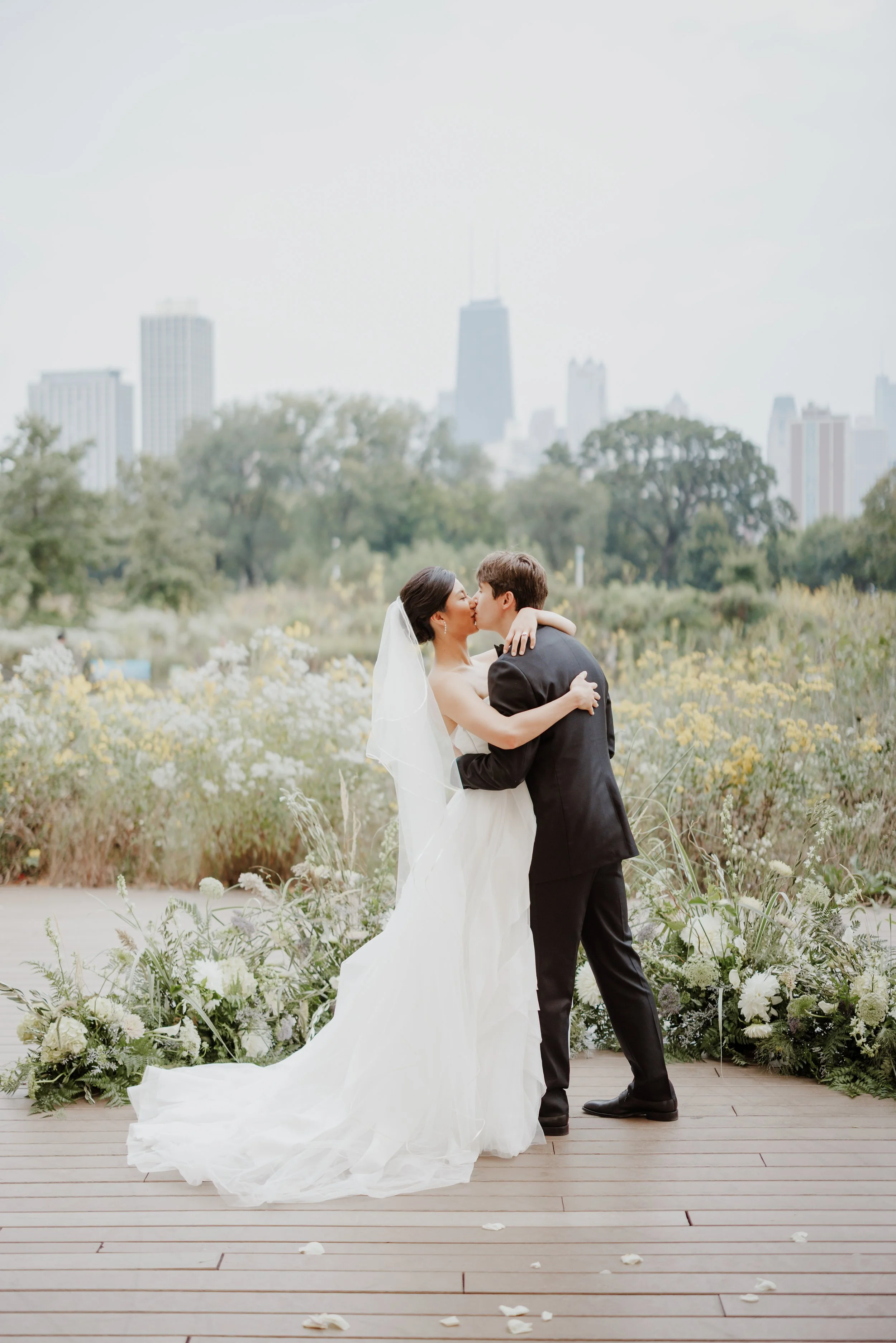 A bride and groom sharing a kiss during their outdoor wedding ceremony, with a city skyline in the background and flowers and greenery decorating the setting.