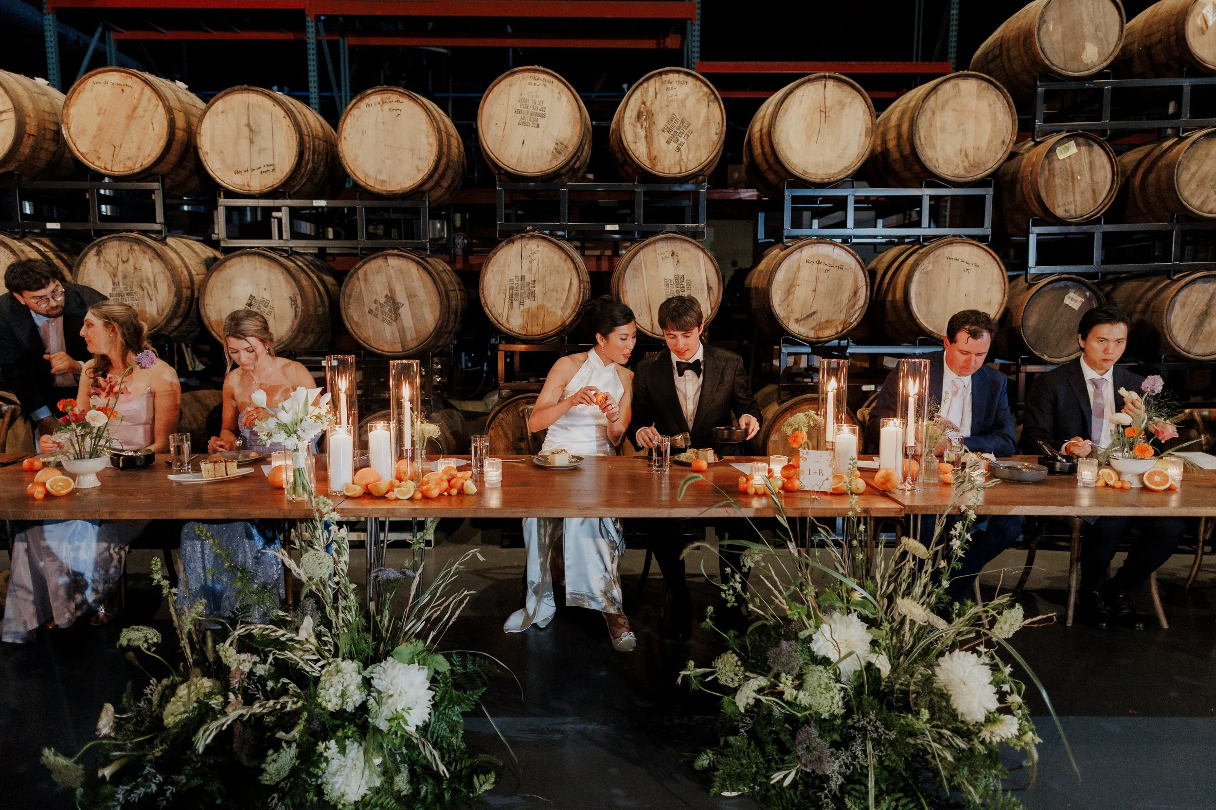 A group of people dressed in formal attire sitting at a long decorated table with flowers and candles, in front of a wall of stacked barrels, in an industrial setting.