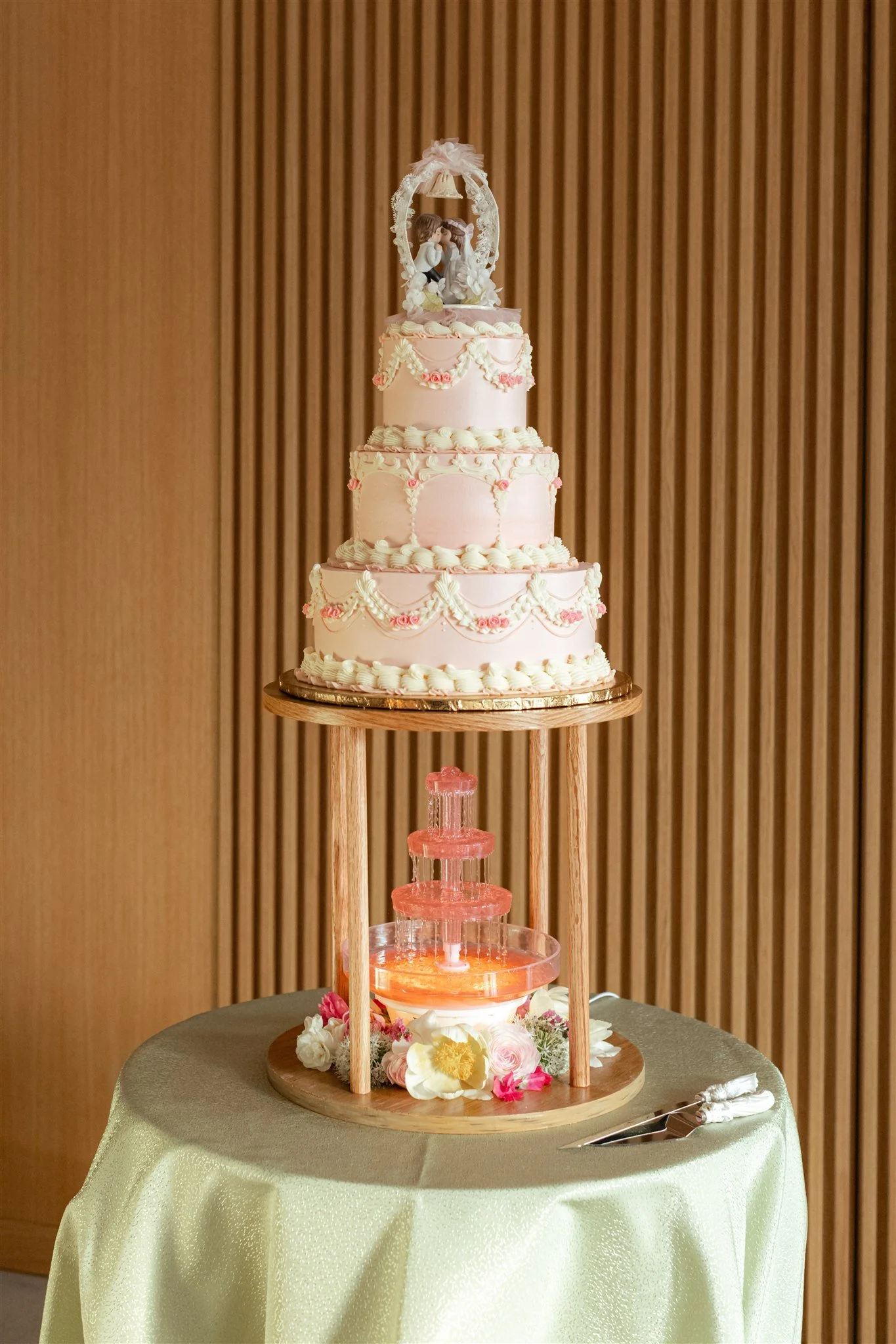 A decorative tiered wedding cake with a cake topper of a bride and groom figurine, set on a round table covered with a beige cloth, with a floral arrangement at the base, in front of a wooden wall.