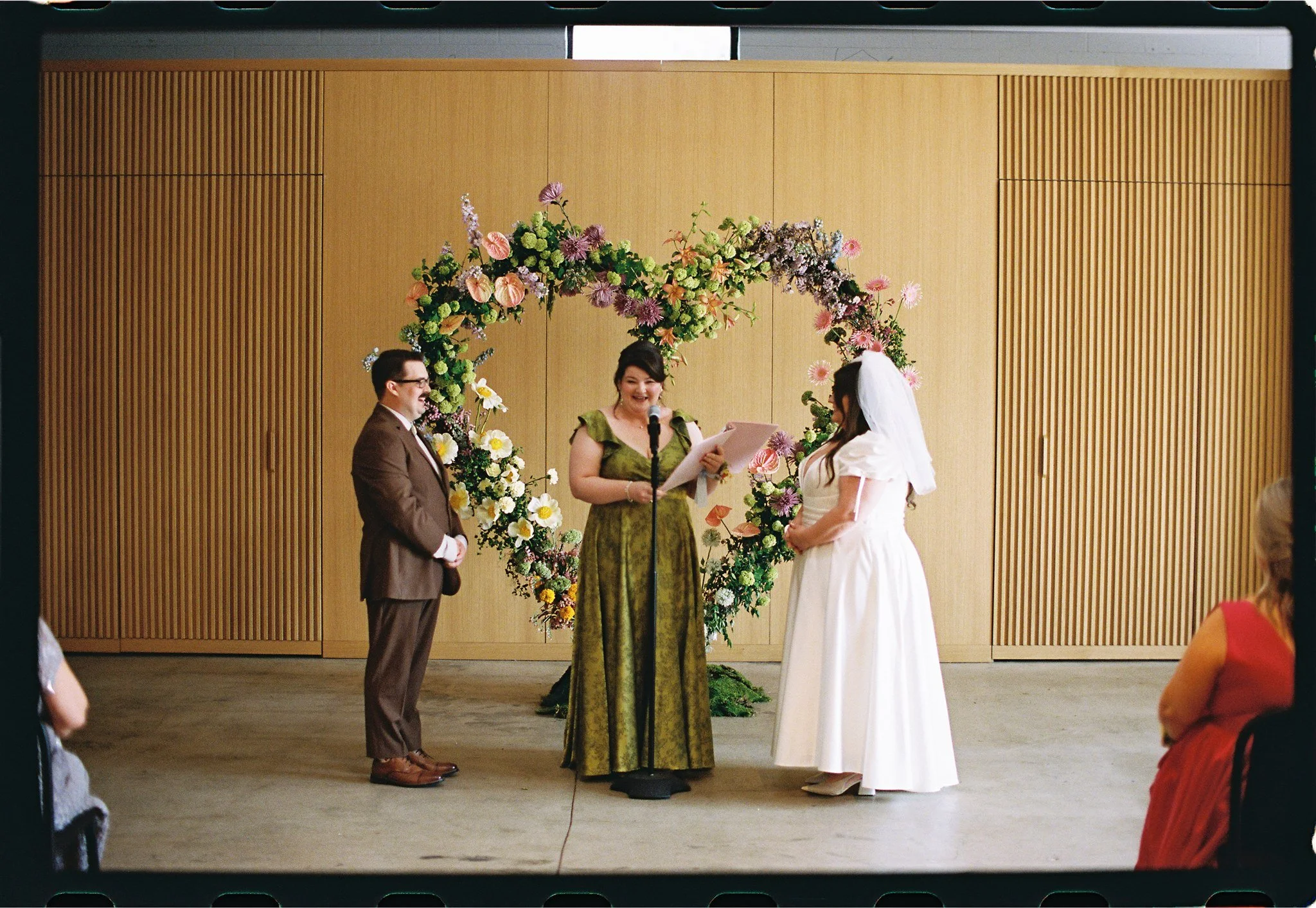 A wedding ceremony with a couple standing before an officiant under a floral arch, with guests seated on either side, in a room with wood-paneled walls.
