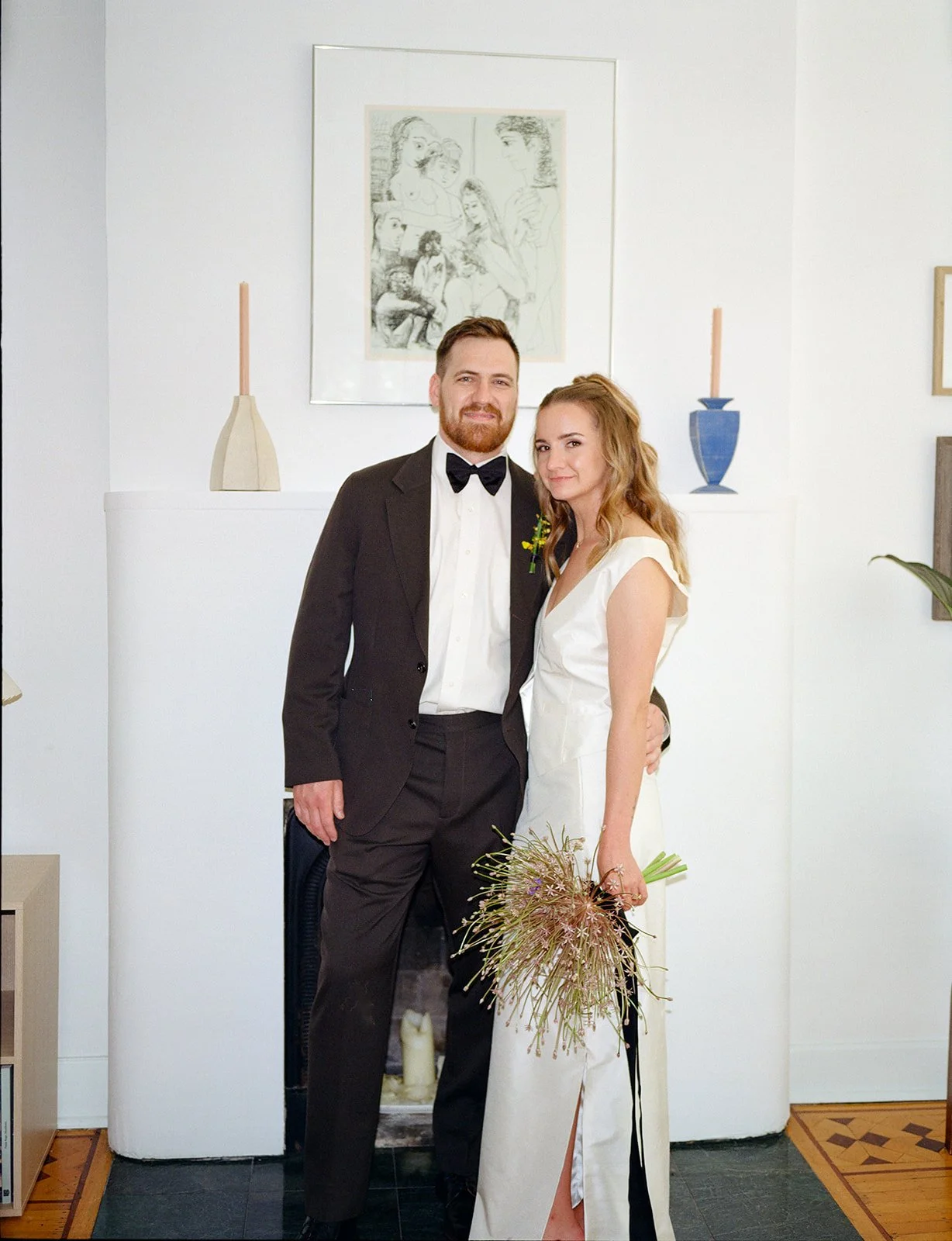 A couple dressed in wedding attire stands closely together in front of a white wall, with a fireplace and framed artwork behind them. The man is wearing a tuxedo with a bow tie, and the woman is in a white gown holding a bouquet of flowers.