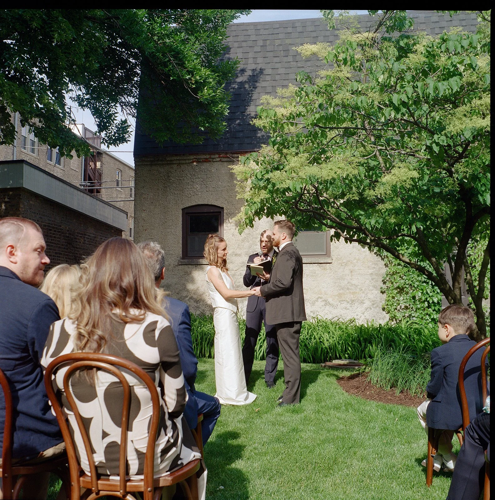 A wedding ceremony taking place outdoors in a garden, with a bride and groom facing each other holding hands, while a person officiates. Guests are seated on wooden chairs, and trees and a building are visible in the background.