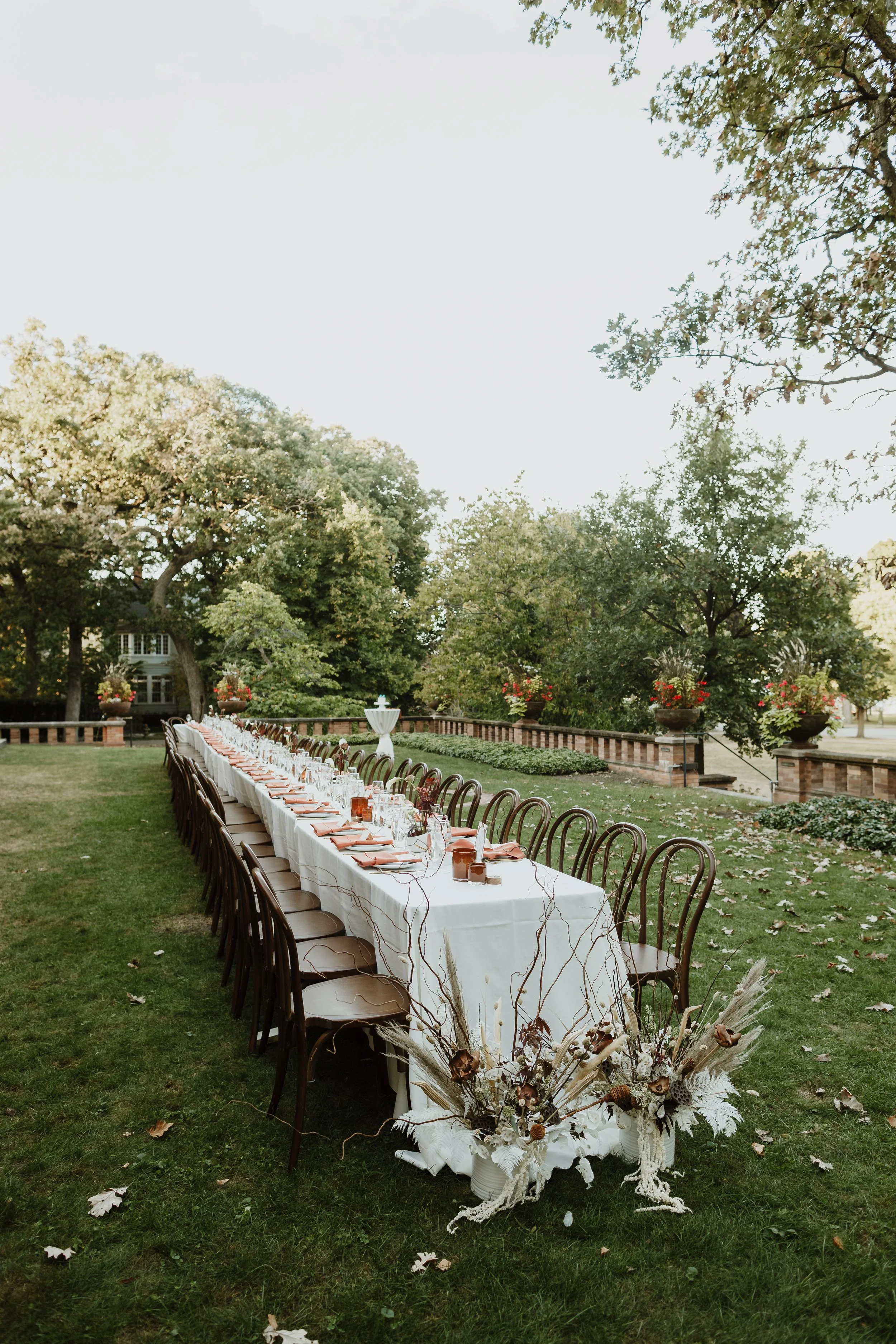 Long banquet table outdoors with white tablecloth, place settings, and floral centerpieces, set in a grassy garden with trees and a brick fence in the background.