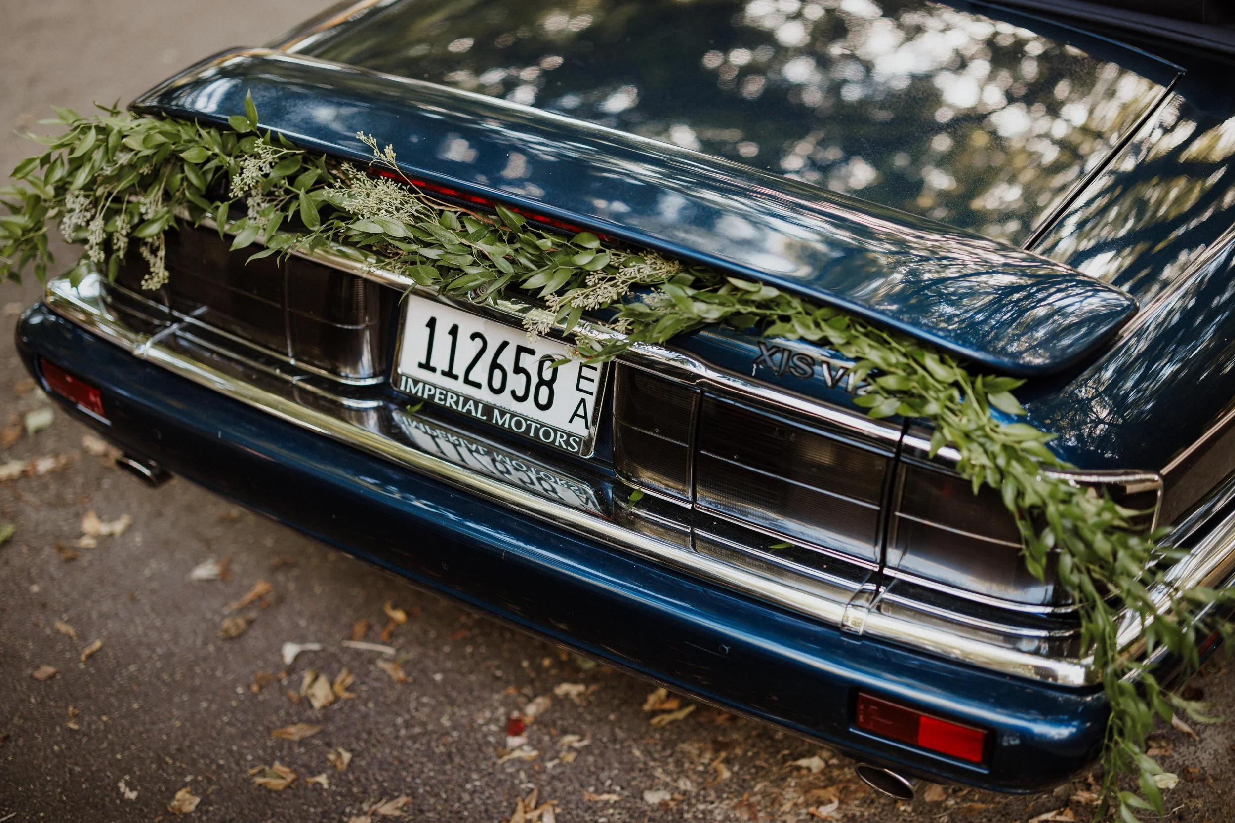 A close-up of the rear of a classic black car decorated with green leaves and white flowers, with a visible license plate reading '112658'.