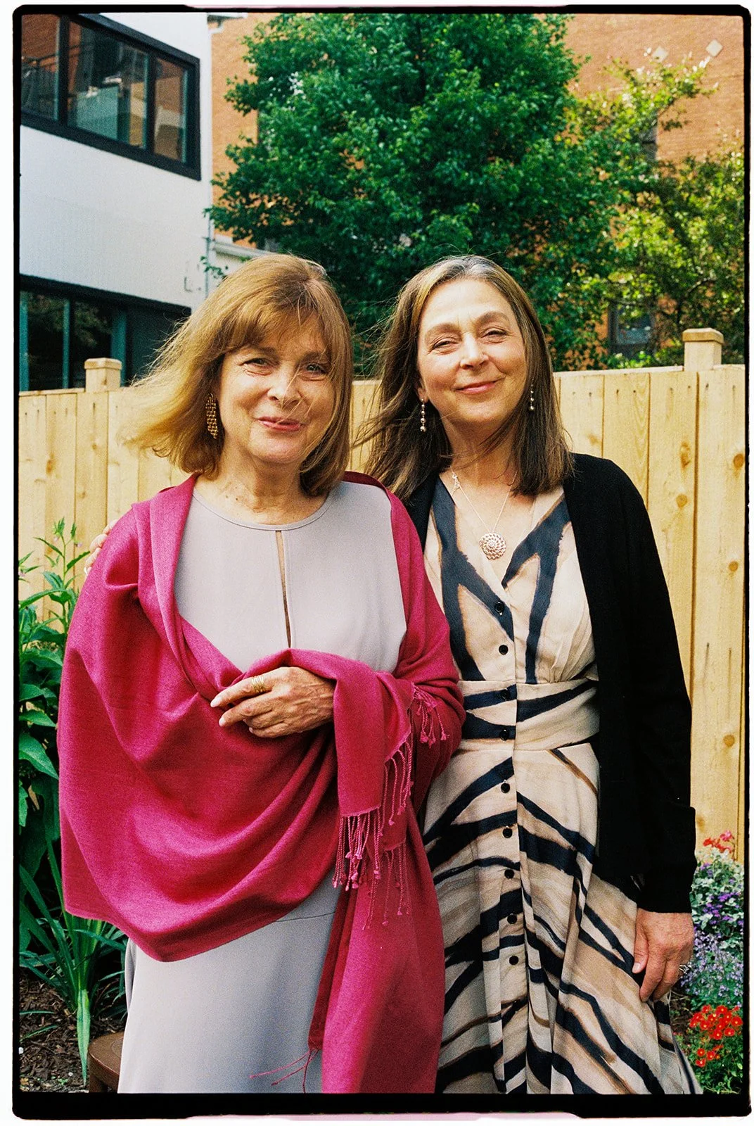 Two women standing outdoors in front of a wooden fence, smiling, with trees and a modern building in the background.