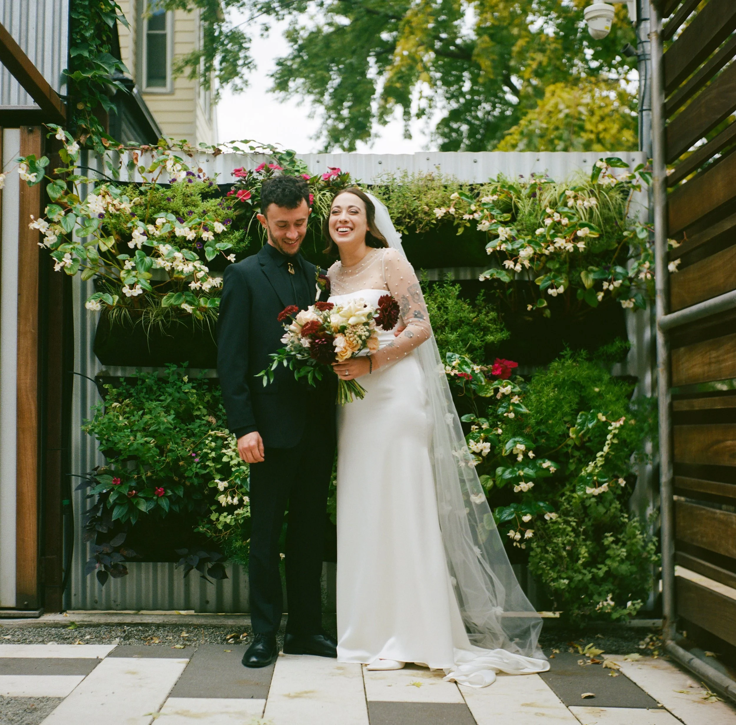 A bride and groom standing together outdoors, smiling. The bride is holding a bouquet of flowers, and the groom is dressed in a dark suit. They are standing behind a low fence surrounded by greenery, with a wooden fence on one side and a garden with flowers behind them.