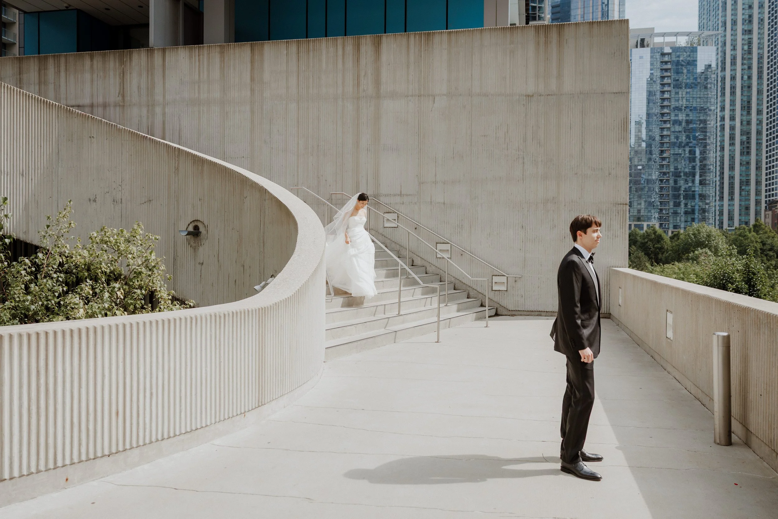 A bride descending a staircase in an urban setting with modern buildings in the background, and a groom standing at the bottom in a tuxedo.
