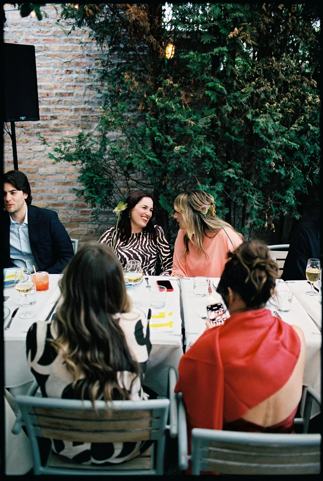 A group of people sitting around a dining table outdoors, smiling and chatting. The setting includes glasses of wine and drinks, with a brick wall and lush greenery in the background, indicating a garden or patio area during a social gathering or cel