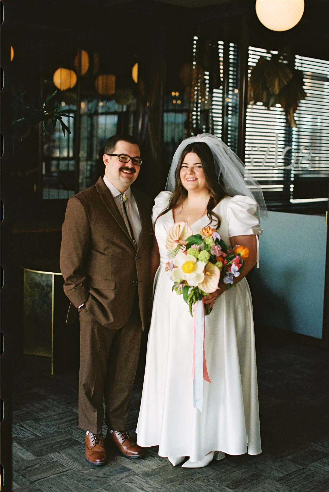 A bride and groom dressed in wedding attire posing indoors. The groom is wearing glasses, a brown suit, and brown shoes. The bride wears a white wedding dress with puffed sleeves, a veil, and holds a bouquet of colorful flowers. The background includ