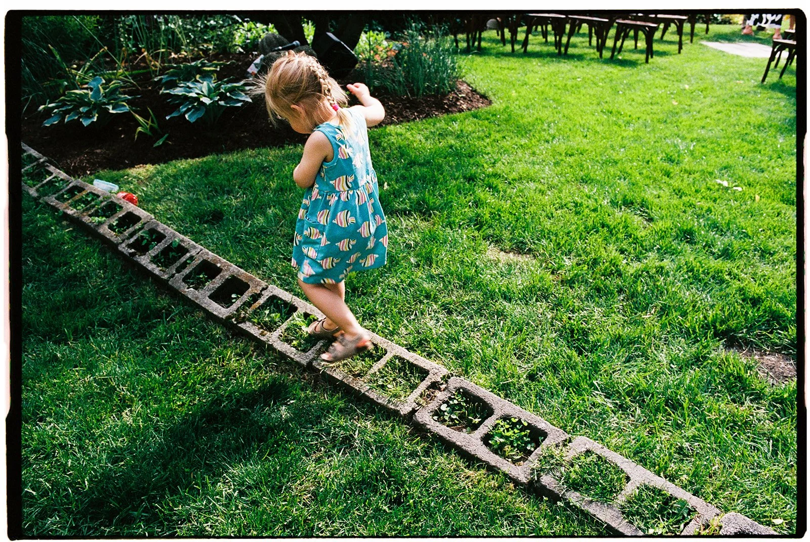 A young girl in a blue dress with colorful patterns walking on a large concrete block placed on the grass in a garden. The garden has green grass, plants, and flowers, with outdoor chairs in the background.