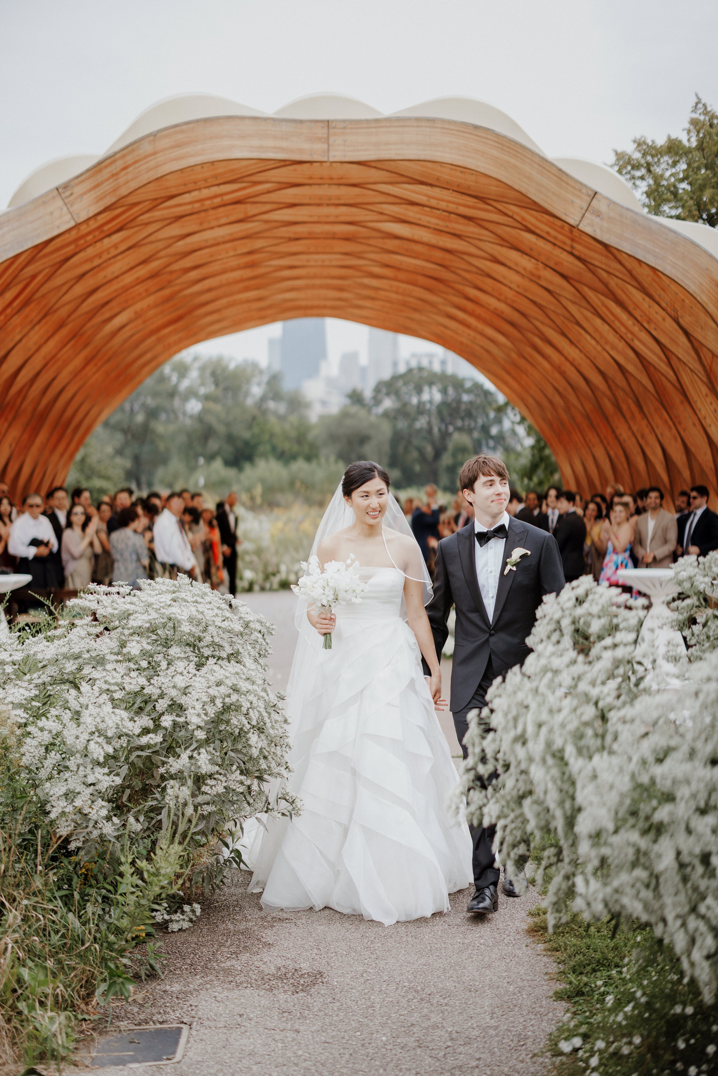 A bride and groom walking hand in hand down a floral aisle at their wedding ceremony, surrounded by guests and under a large wooden arch.