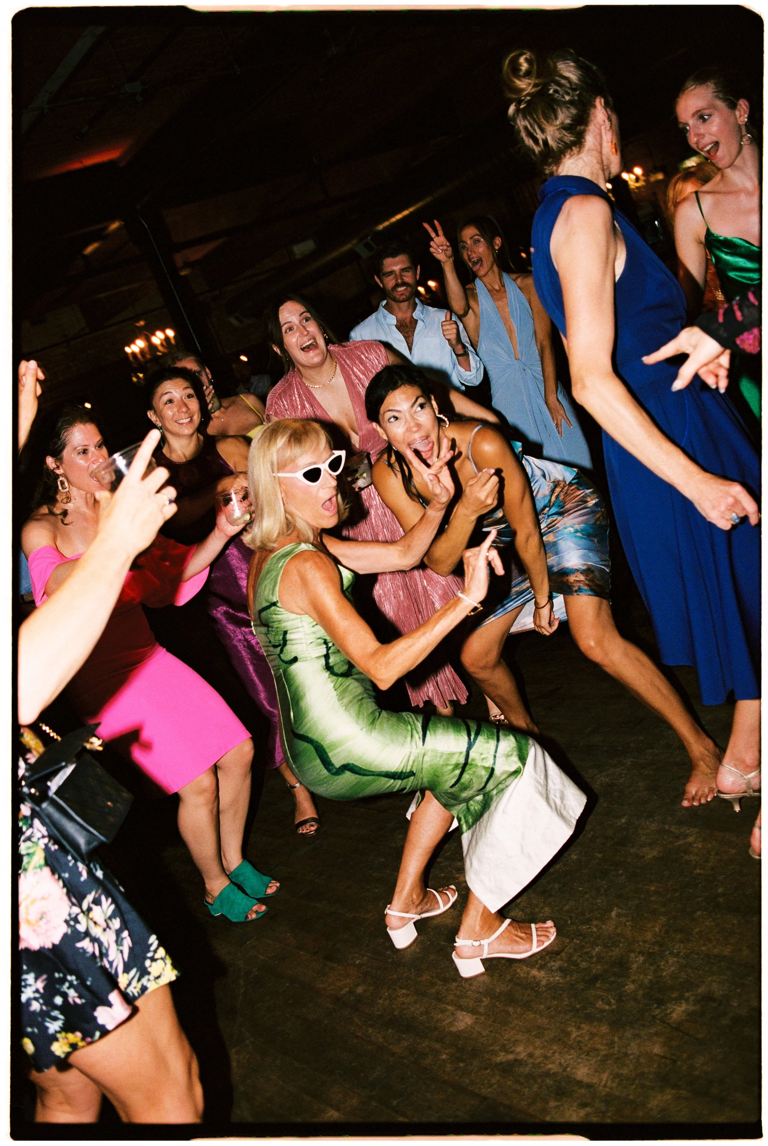 A group of people dancing and having fun at a party or celebration, with some women wearing colorful dresses and heels, and one woman in a green dress with white heels. They are indoors with a dark ceiling and warm lighting in the background.