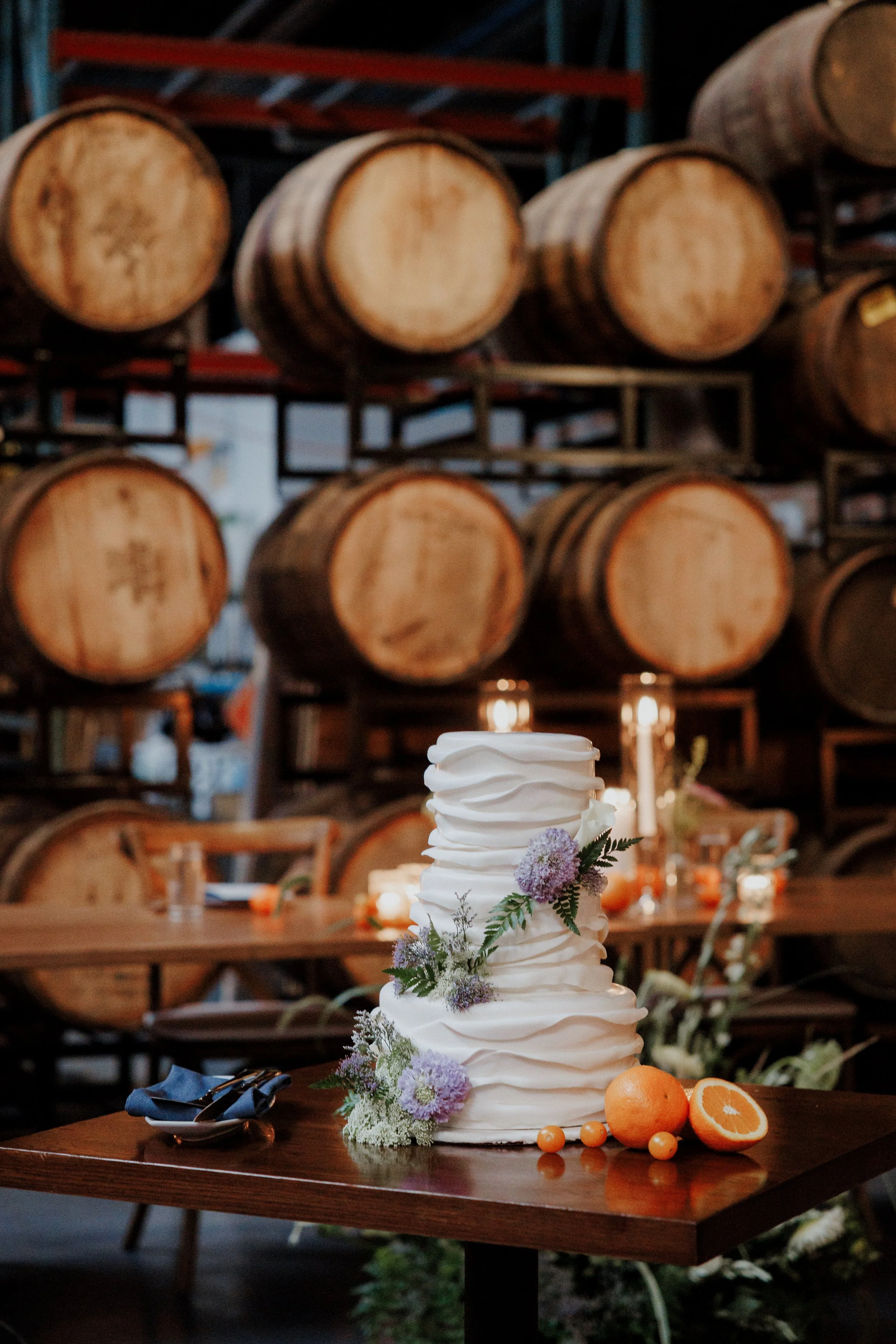 A multi-tiered white wedding cake decorated with purple and white flowers, placed on a wooden table, with oranges and orange slices nearby, in a rustic setting with barrels in the background.