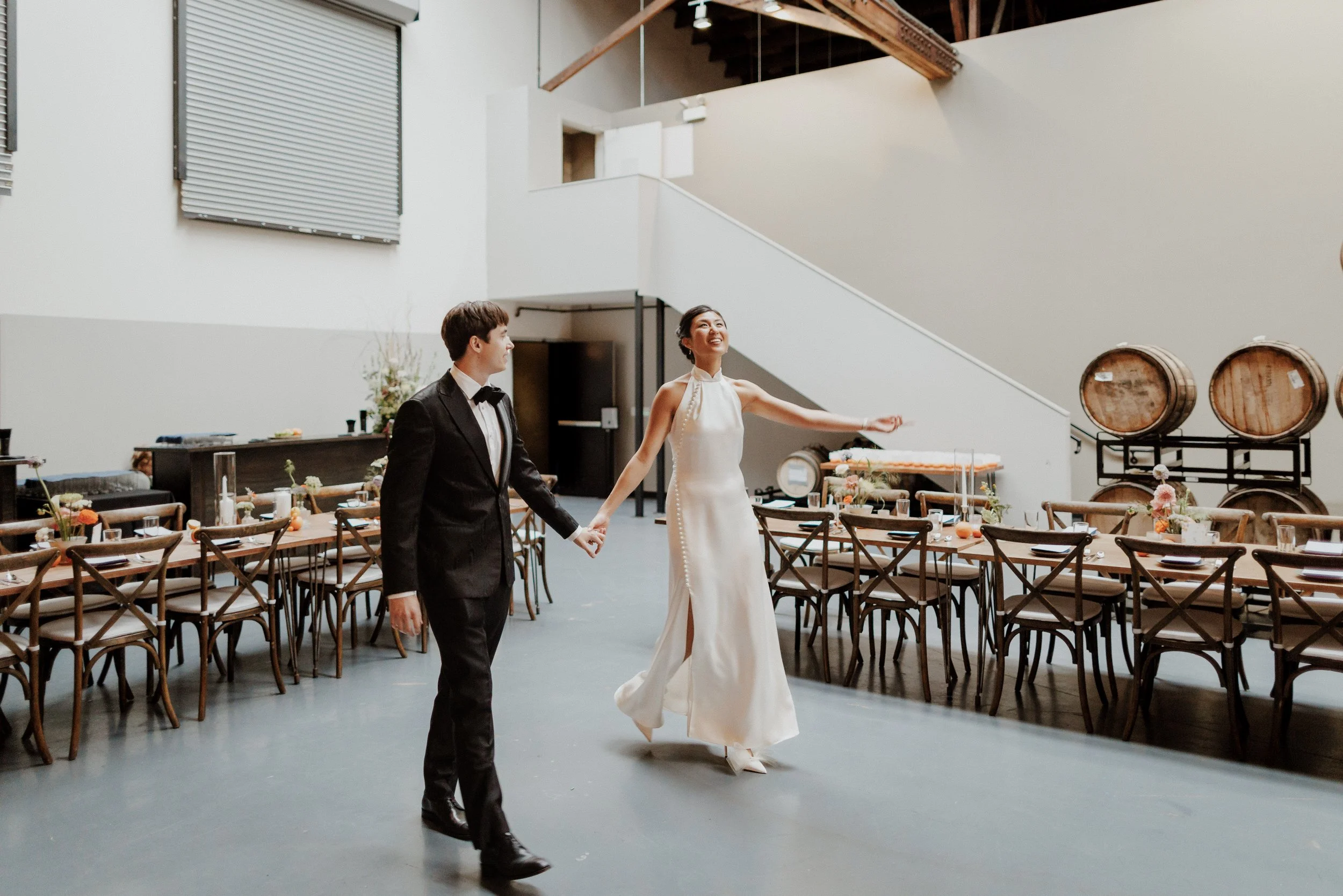 A bride and groom holding hands inside a rustic wedding venue with wooden barrel decorations and arranged tables.