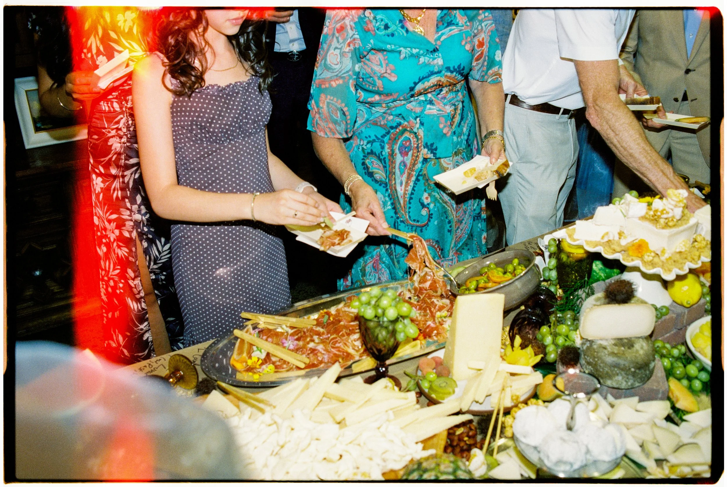 People serving themselves food at a buffet table with various cheeses, fruits, and appetizers.