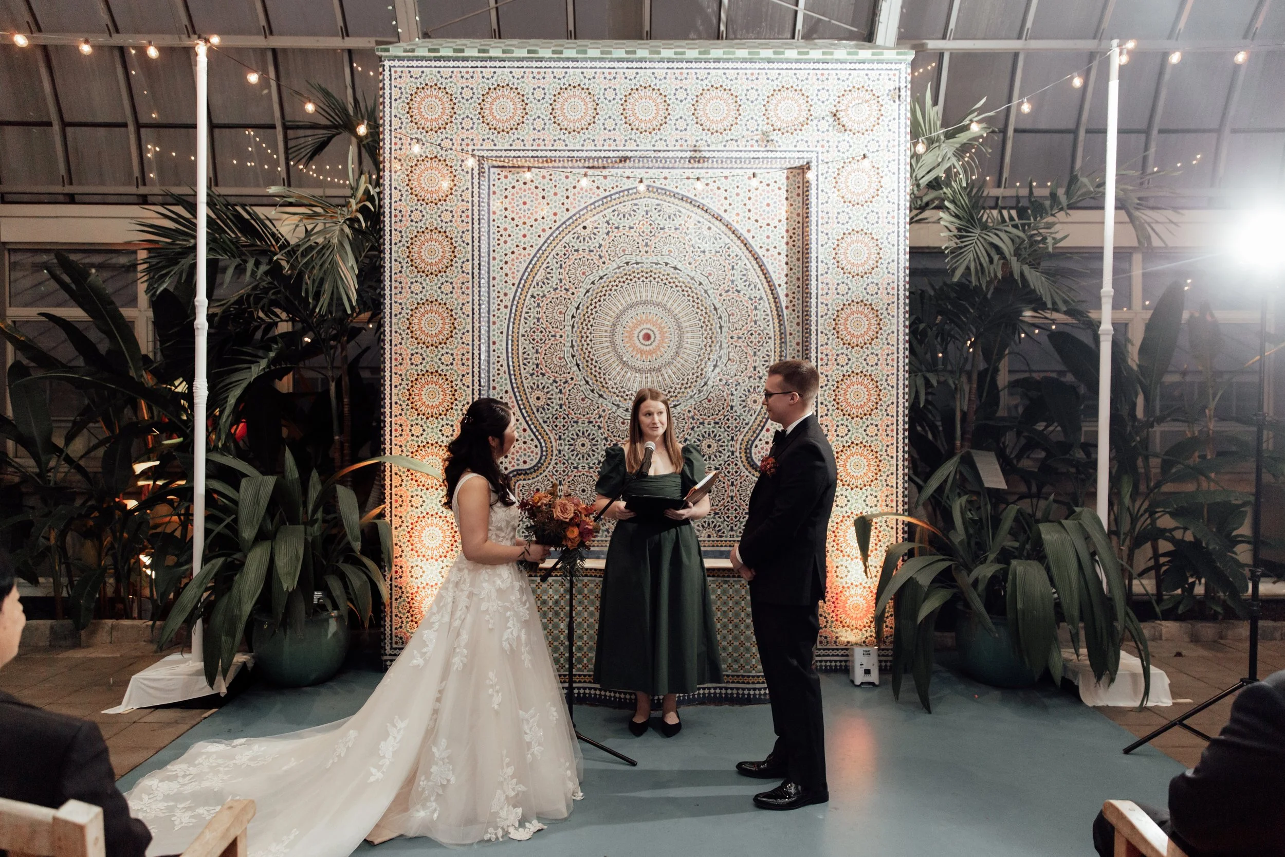 A wedding ceremony with a bride and groom standing before an officiant inside a glass-walled venue decorated with string lights and large potted plants, with a patterned backdrop behind them.