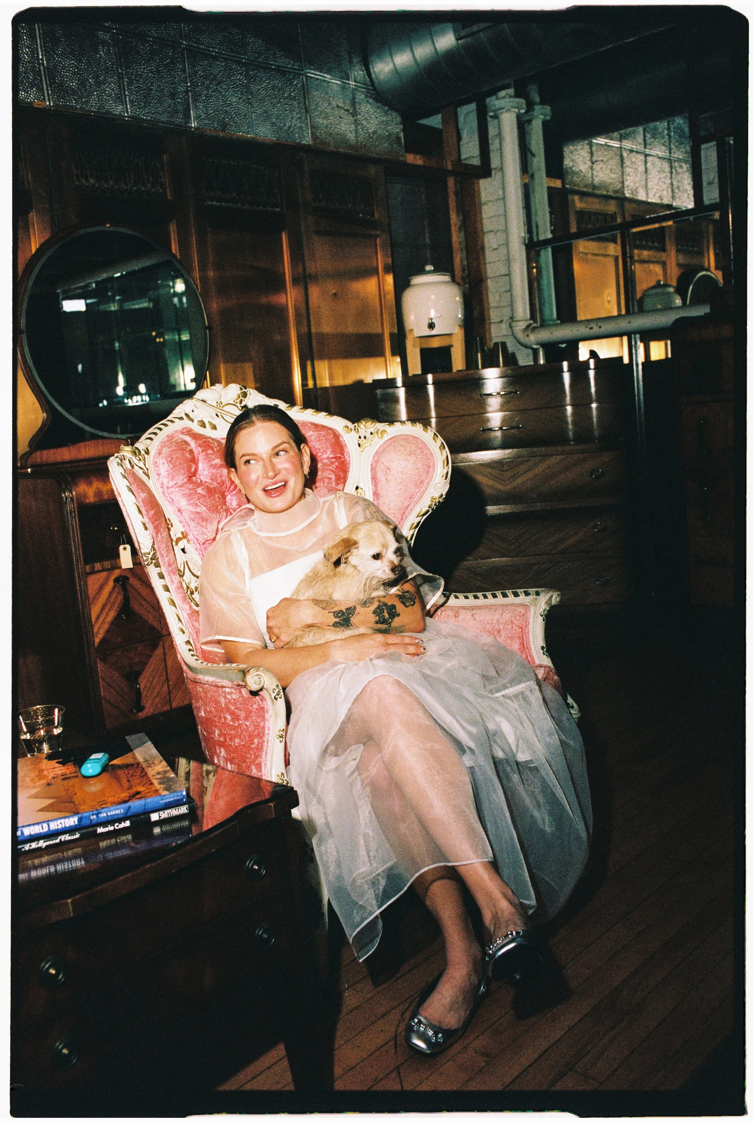 A woman wearing a sheer, off-white dress with tattoos on her arms, sitting on an ornate pink and gold armchair, holding a small dog. She is smiling and looking to her left in a dimly lit room with wooden flooring, a dark wooden dresser with a stack o