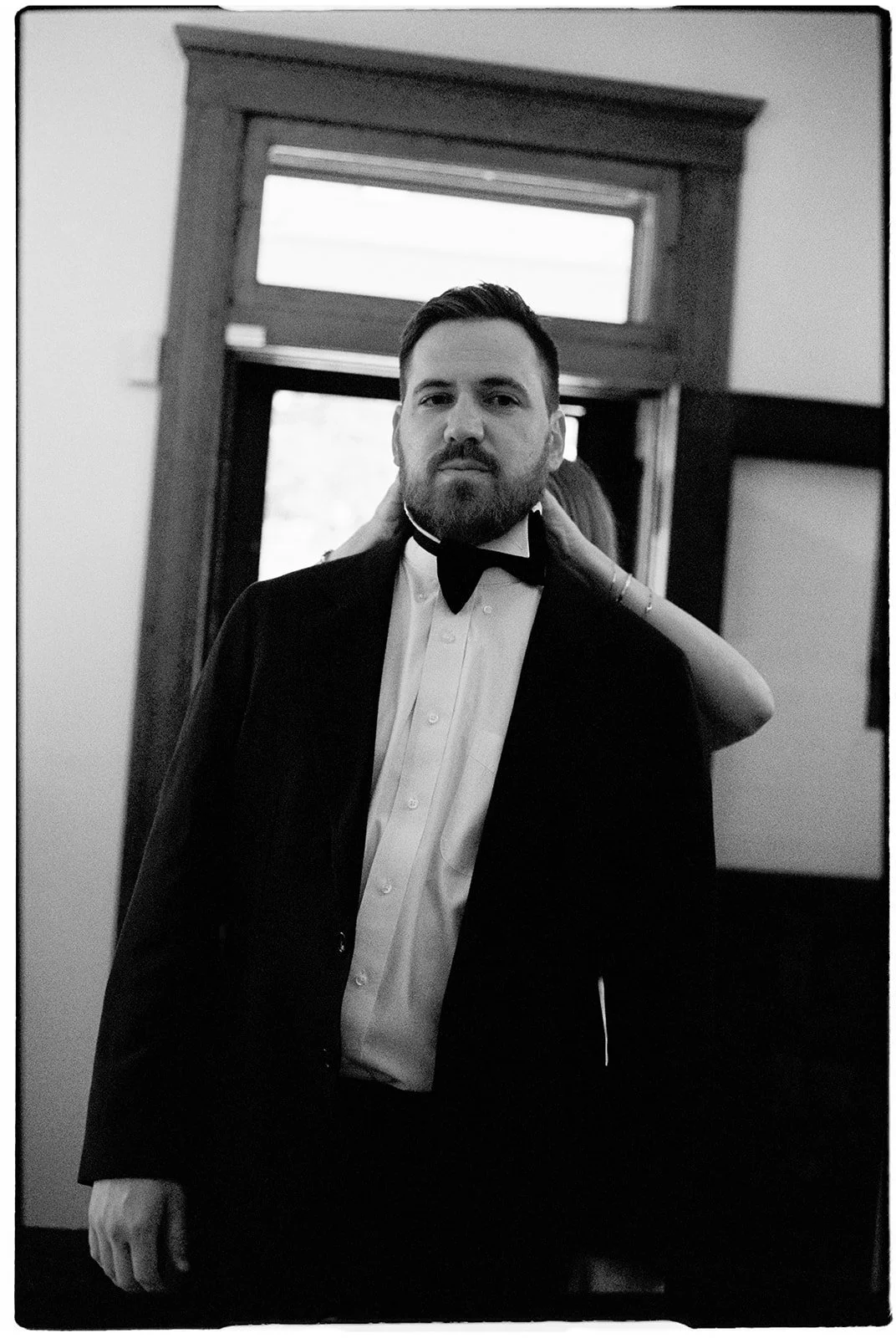 A man with a beard in formal attire, including a tuxedo and bow tie, standing indoors in front of a window, while a person behind him adjusts his collar.