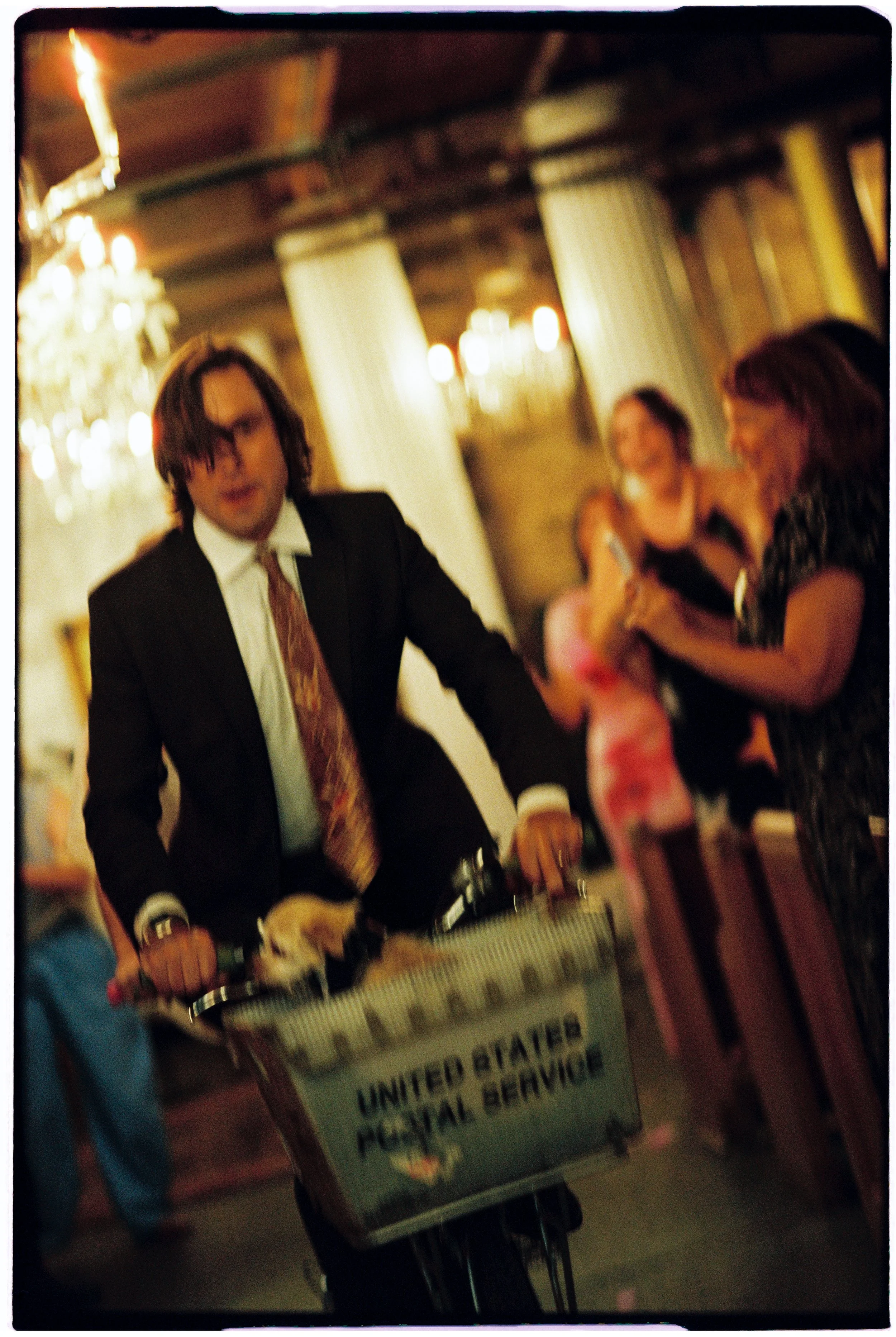 A man in a suit and tie pushing a cart with a United States Postal Service box, with two women in the background having a conversation in a warmly lit room.