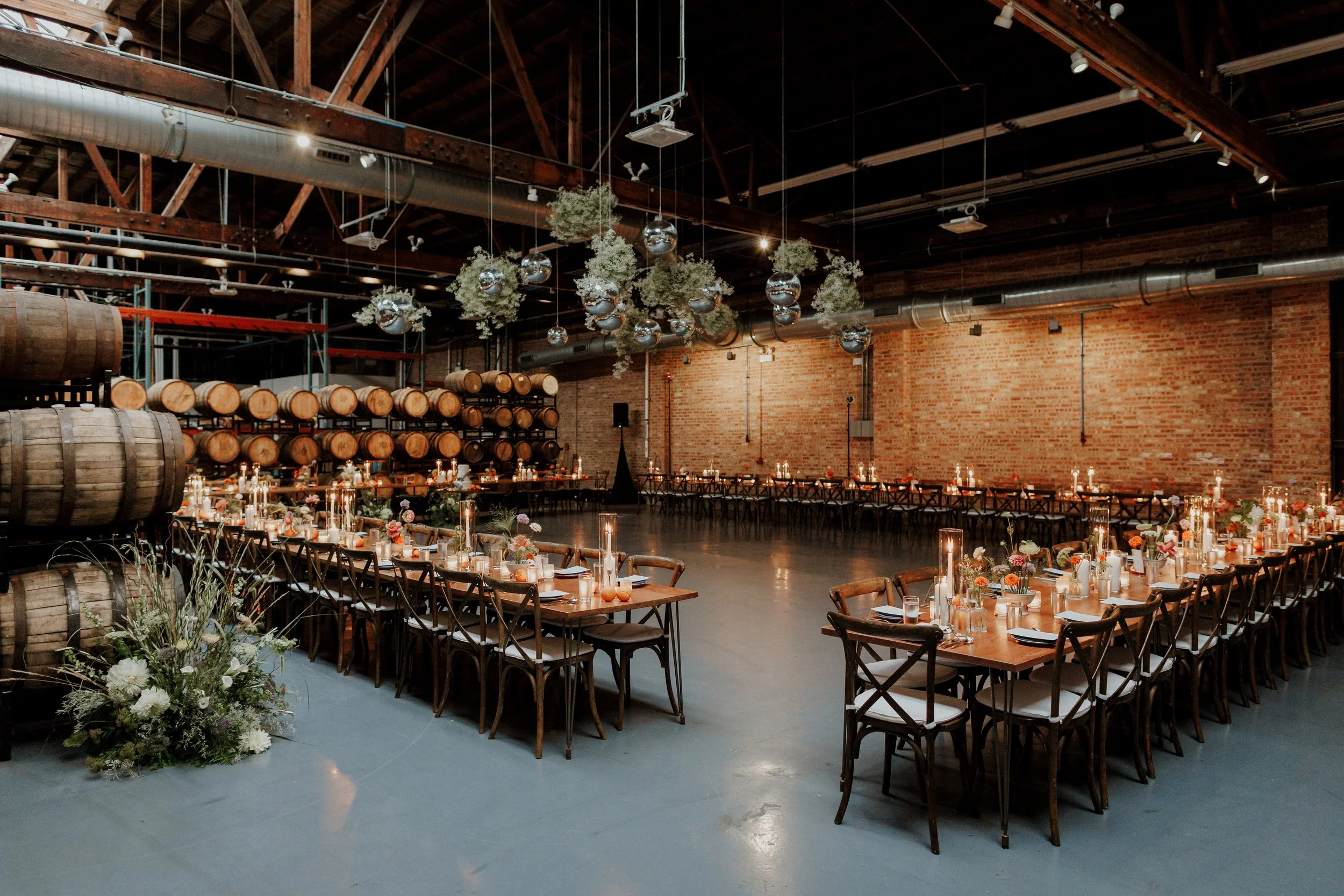 Interior of a rustic event space with wooden beams and exposed brick walls, decorated with hanging greenery and reflective ornaments, set up for a celebration with tables, candles, and floral arrangements.