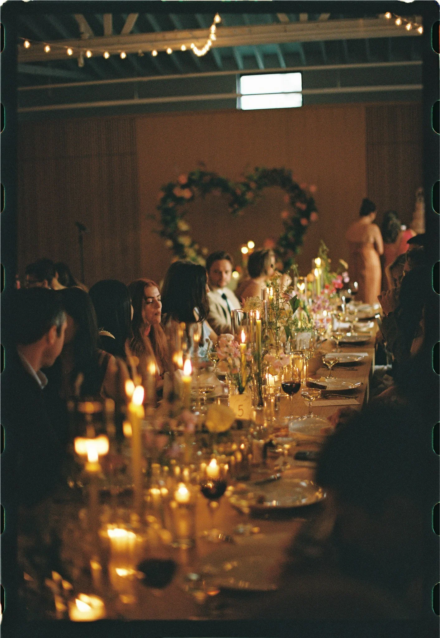 A dimly lit indoor wedding reception with a long rectangular table decorated with candles and flowers. Guests are seated on both sides of the table, and a floral heart-shaped wedding arch is visible in the background. String lights hang from the ceil
