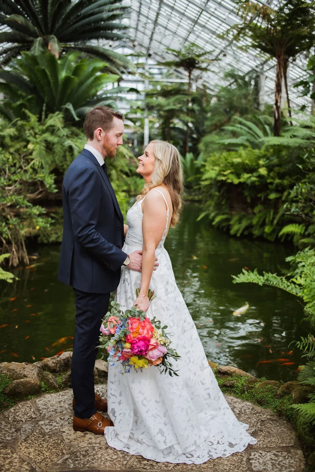 Couple in wedding attire holding hands and smiling at each other in a lush, green indoor garden with a pond and koi fish.