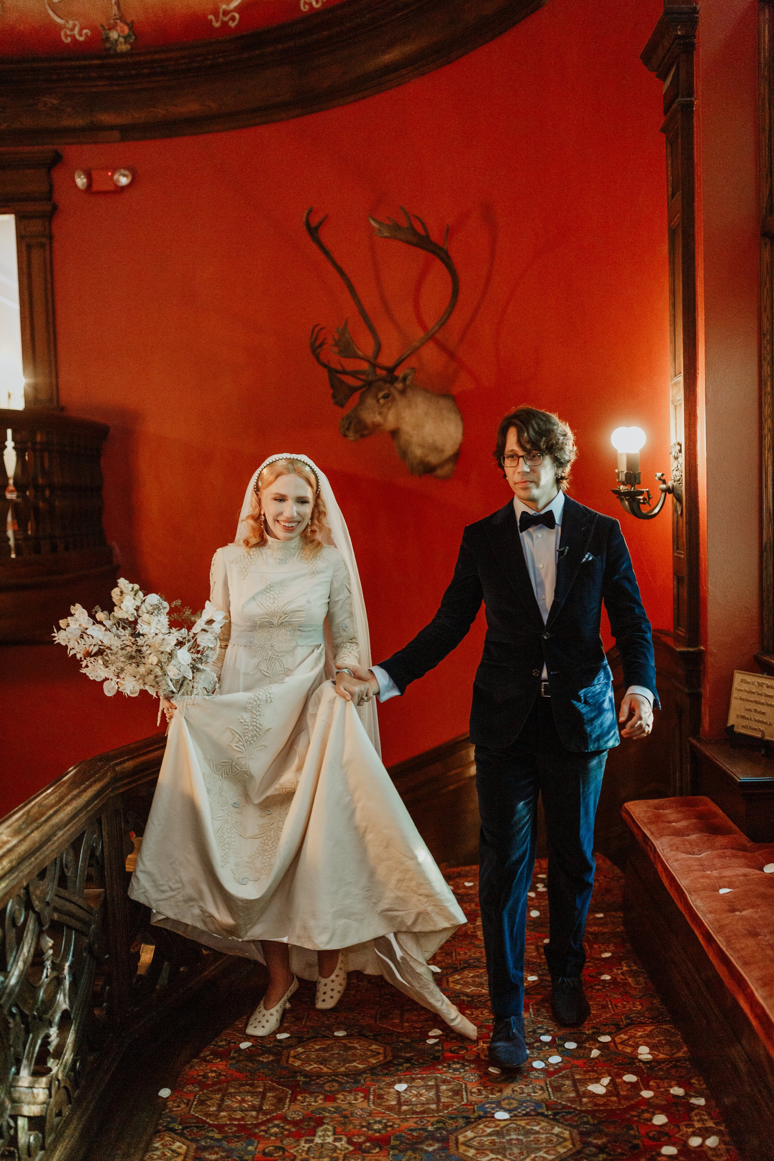 A bride in a white wedding dress holding a bouquet, and a groom in a dark tuxedo, walking down a staircase holding hands, in an elegant room with a mounted moose head on a red wall in the background, and scattered confetti on the floor.