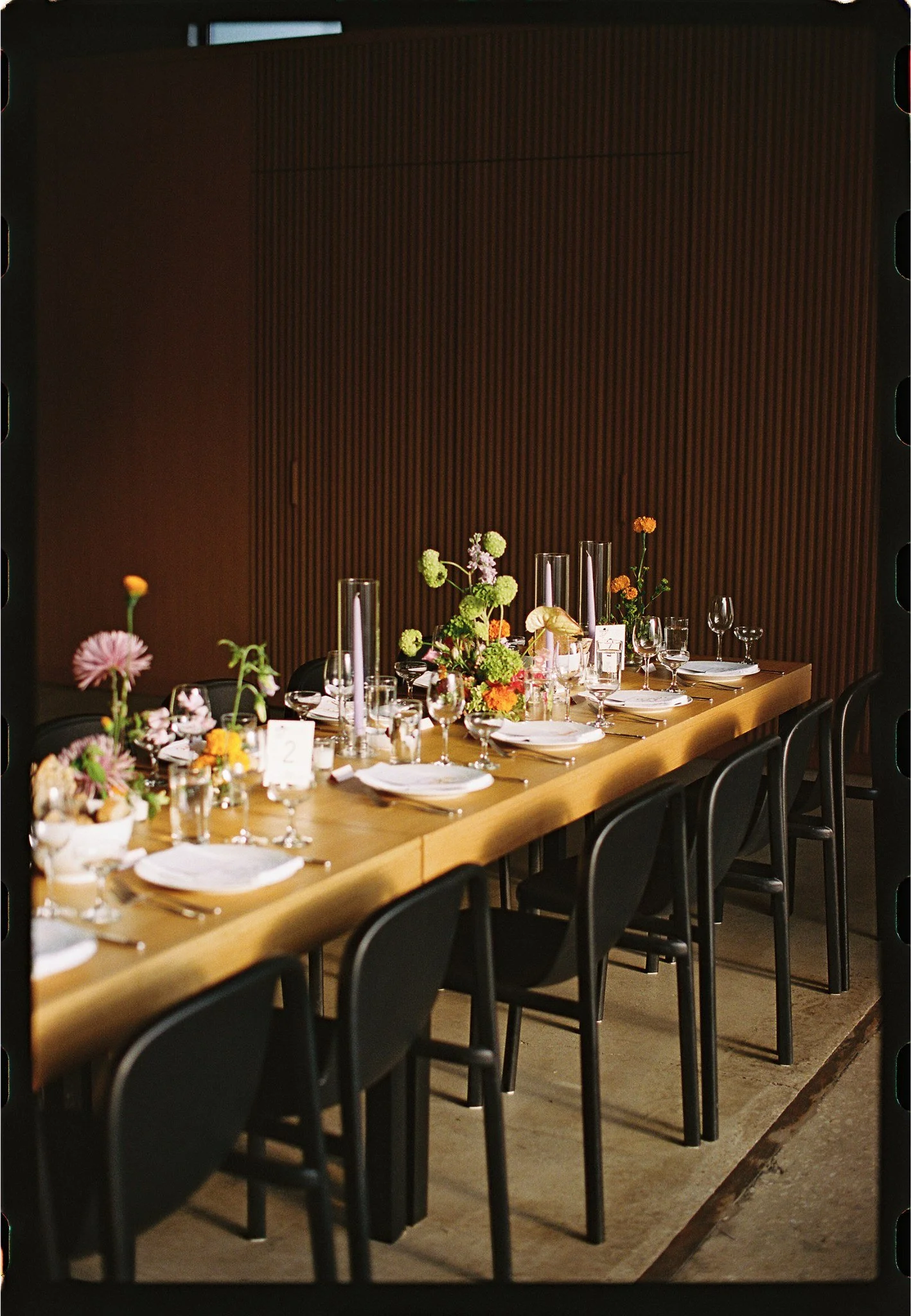 A long wooden table decorated with colorful flowers and candles, set for a formal dinner with plates, glasses, and napkins, against a dark wooden wall background.
