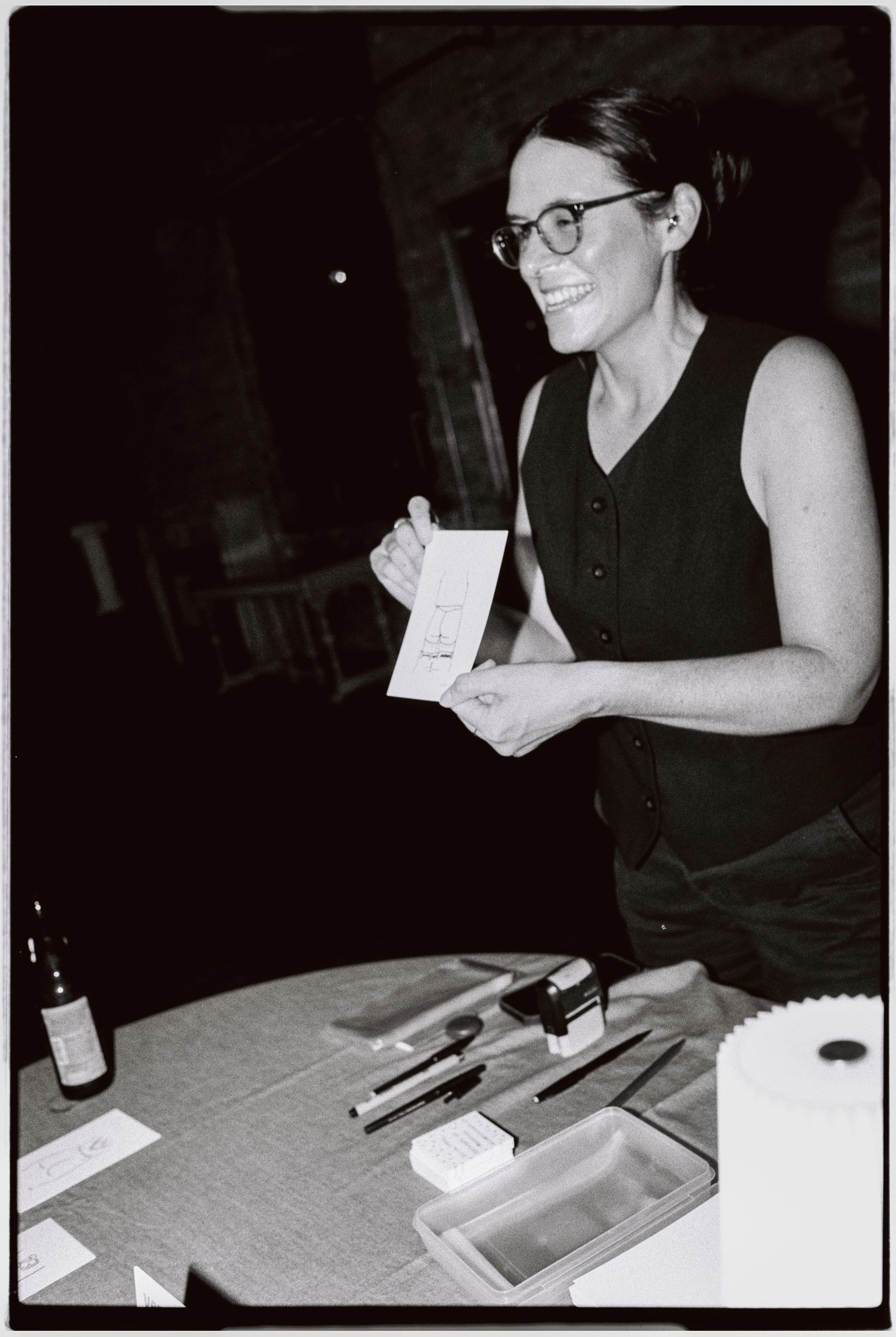 A woman with glasses smiling and holding a piece of paper, standing behind a table with various office supplies, including pens, a stamp, paper, and a plastic container. The setting appears to be an office or workspace.
