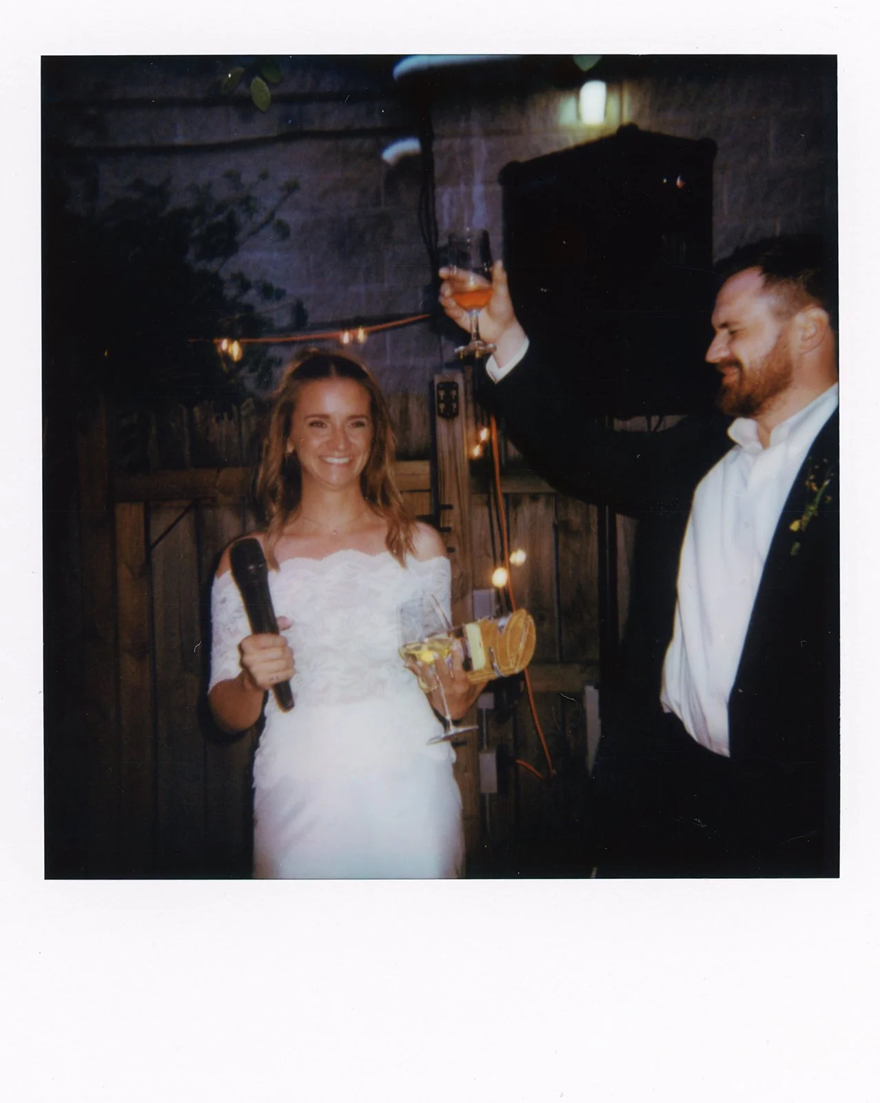A bride holding a microphone and a glass of champagne, smiling, while a man in a tuxedo raises a glass in a toast, celebrating at a wedding reception.