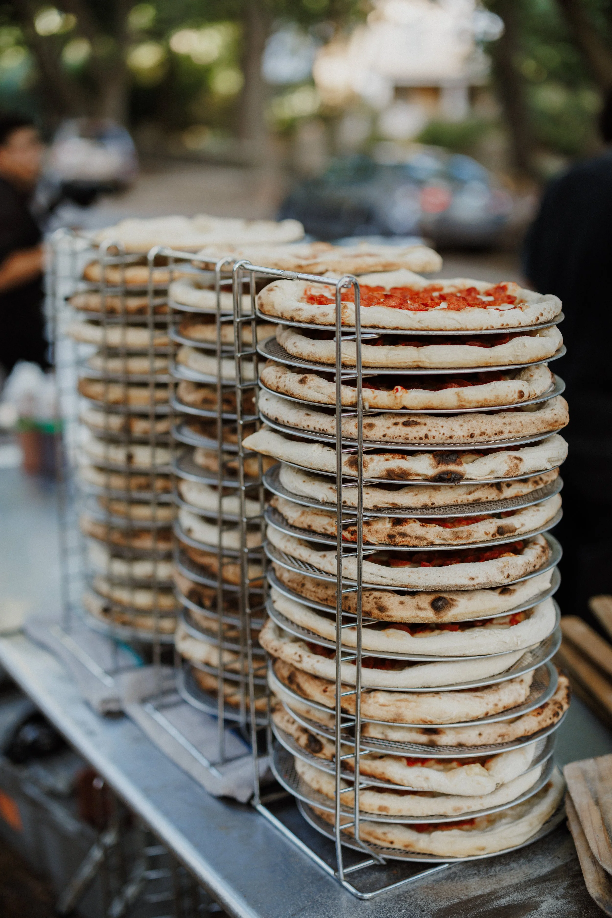 Multiple pizzas with toppings on metal racks outdoors, with blurred background of trees and people.