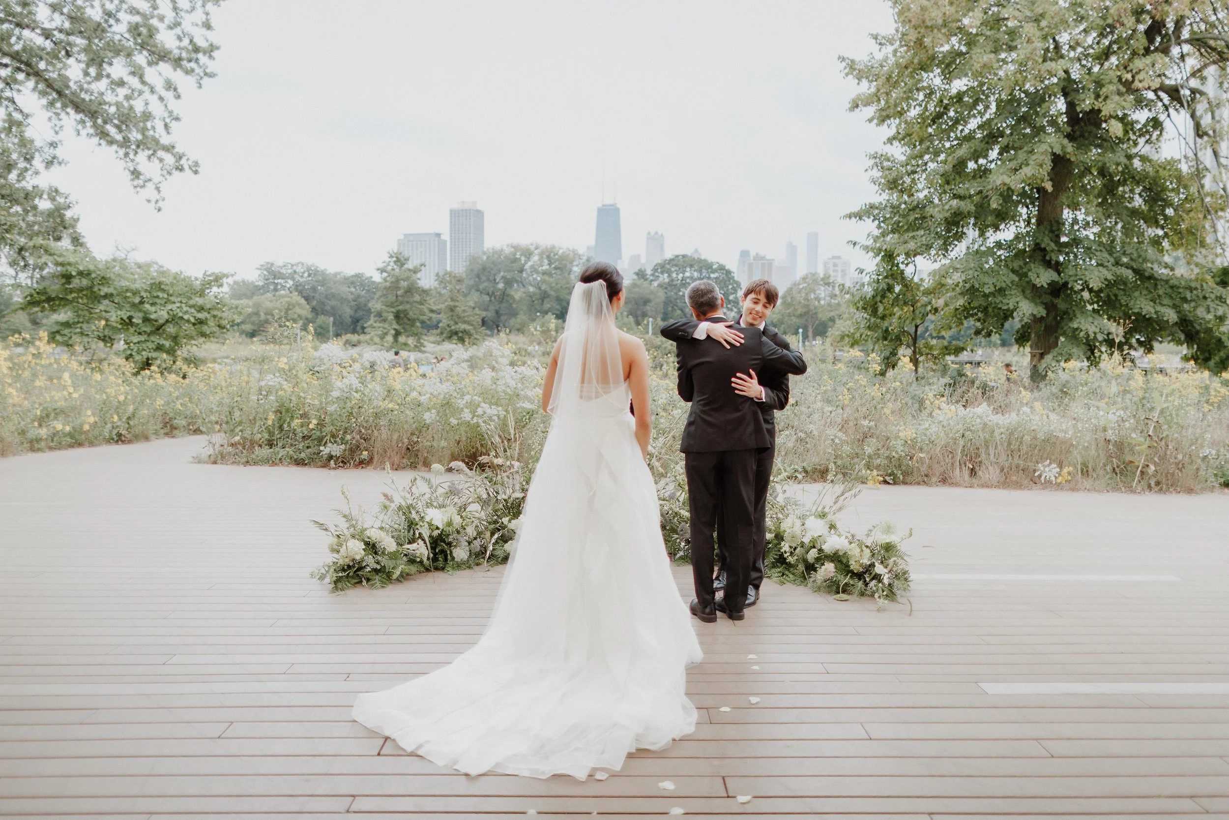 A bride and groom embracing during their wedding ceremony outdoors, with the city skyline visible in the background.