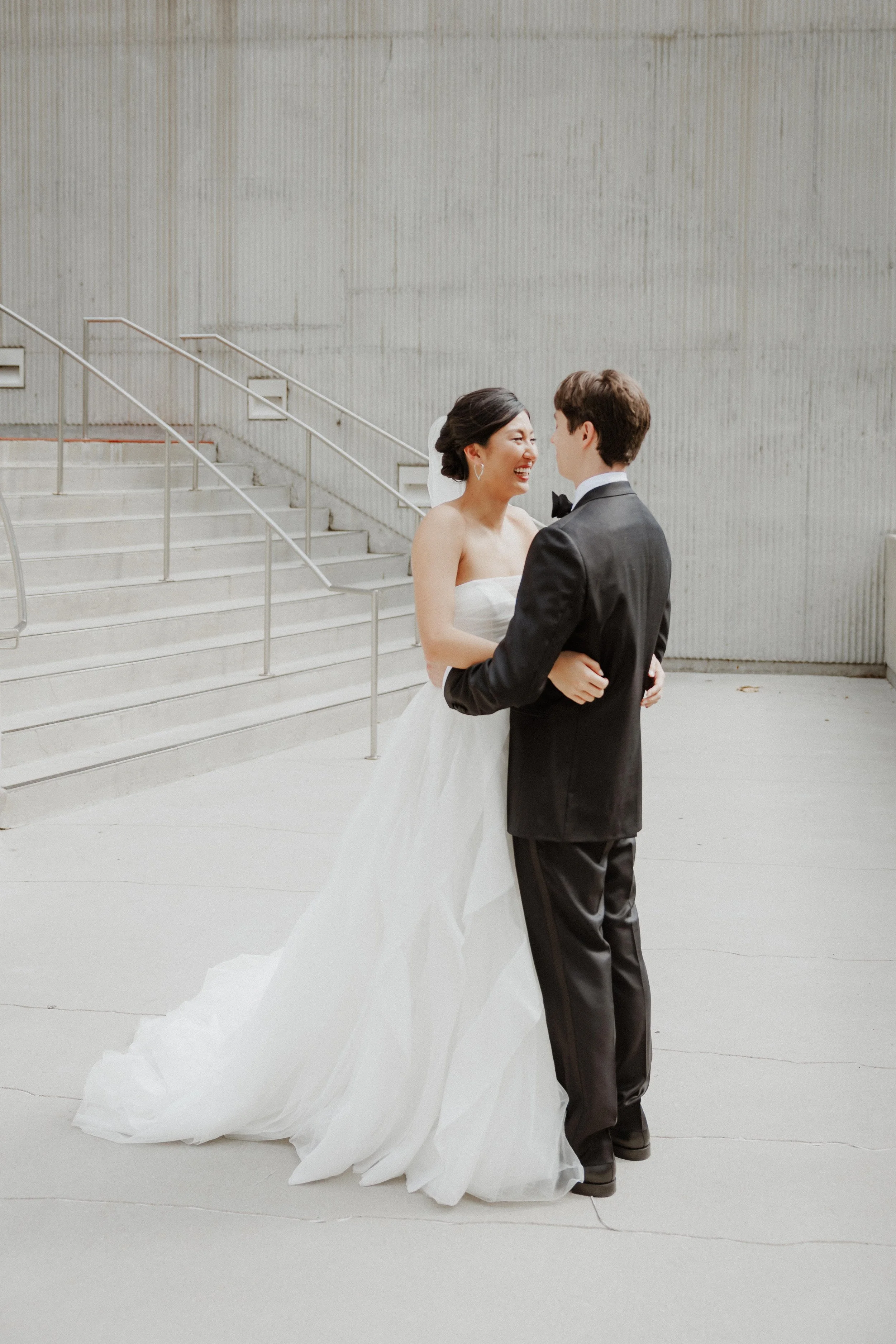 A bride and groom dancing together in an outdoor, modern setting with concrete stairs and metal railings, the bride wearing a strapless wedding gown and the groom in a black tuxedo.